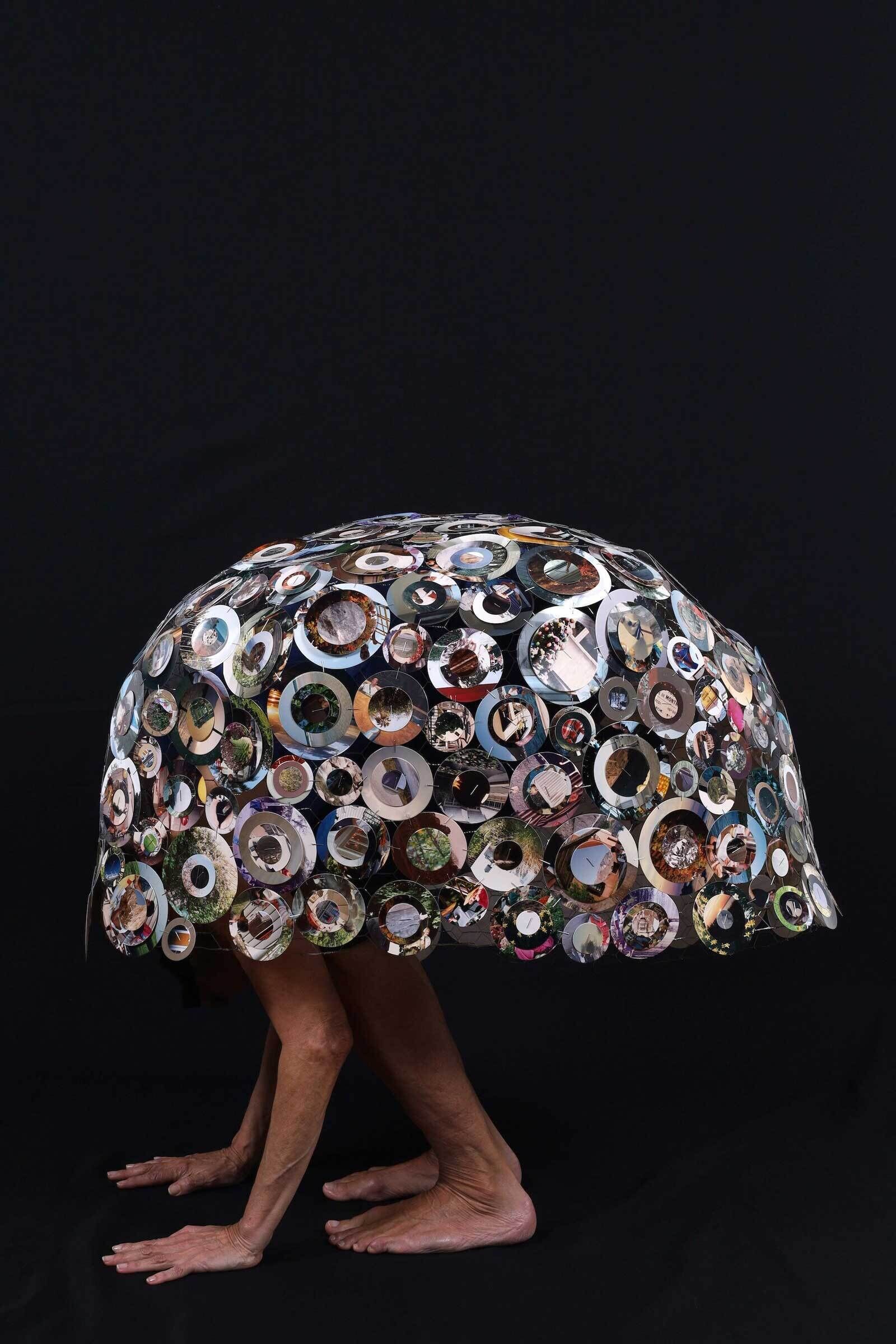 A person is bent over with a dome covered in circular photos and mirrors resting on their back.