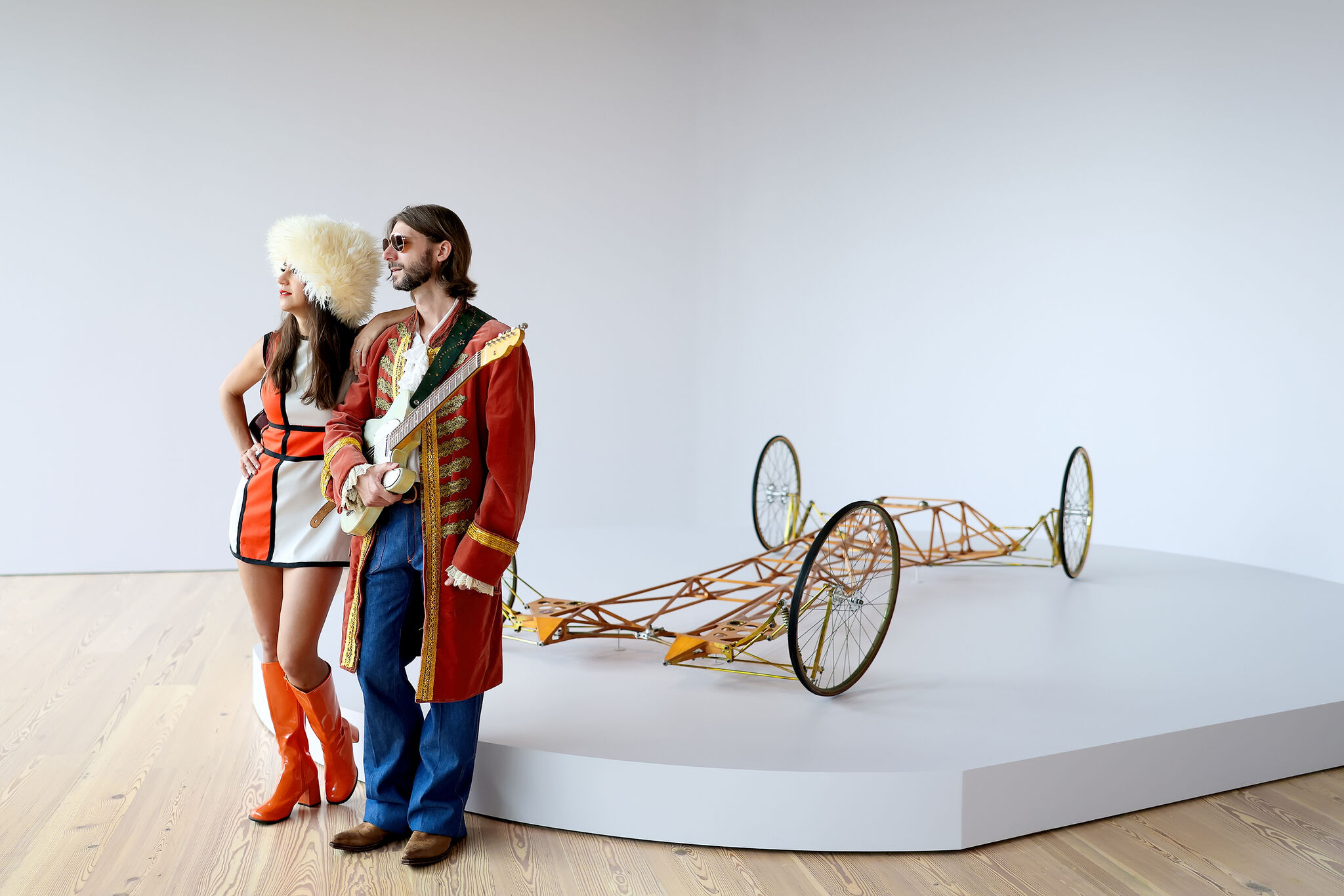 Two people in colorful retro outfits stand together; one holds a guitar. A wire-frame vehicle is displayed behind them.