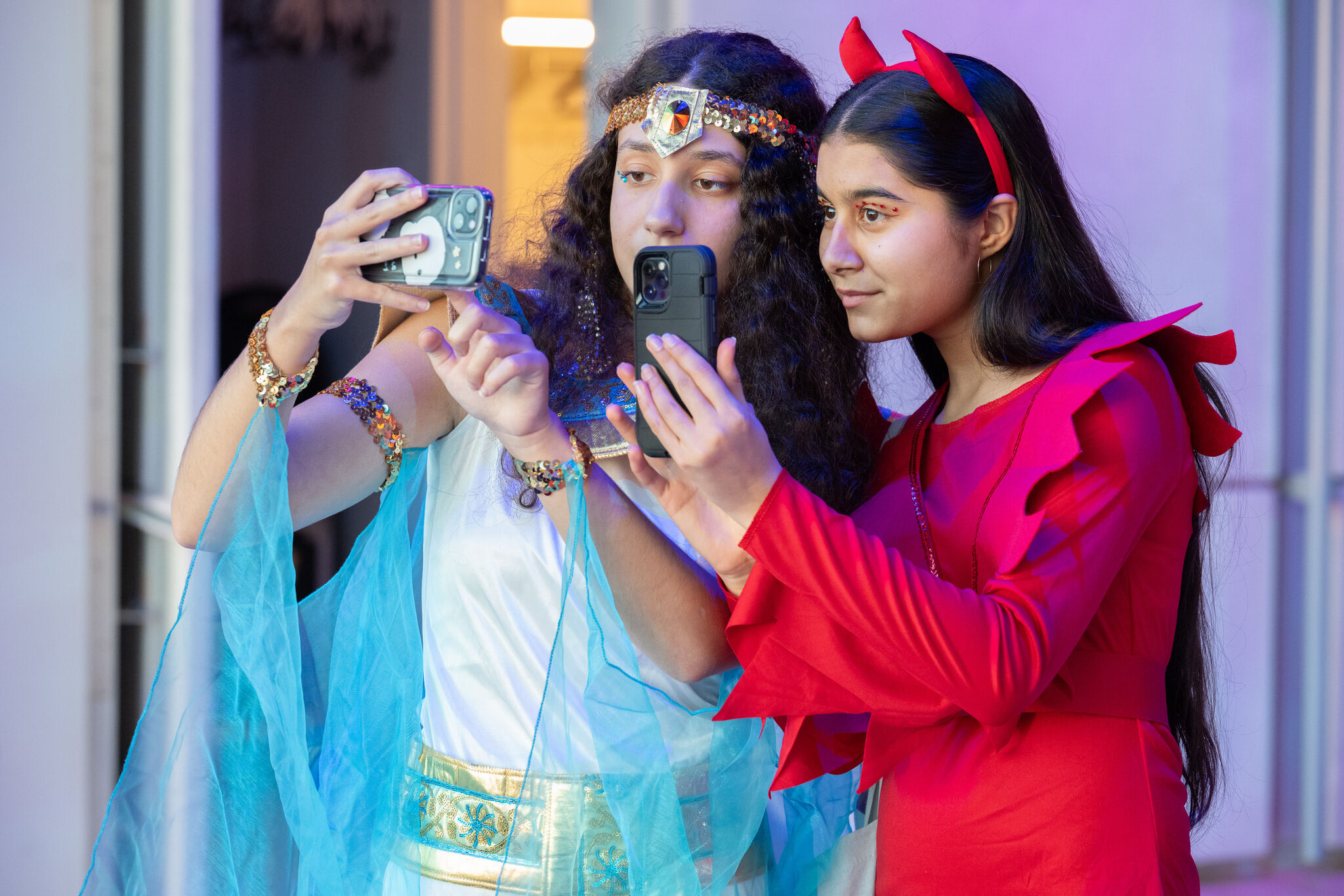 Two girls in costumes, one as a goddess and one as a devil, taking selfies together with their phones.