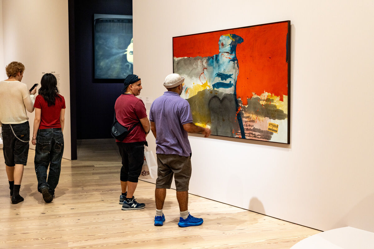 Four people in a museum, two closely observing a large abstract painting with bright red and blue colors.