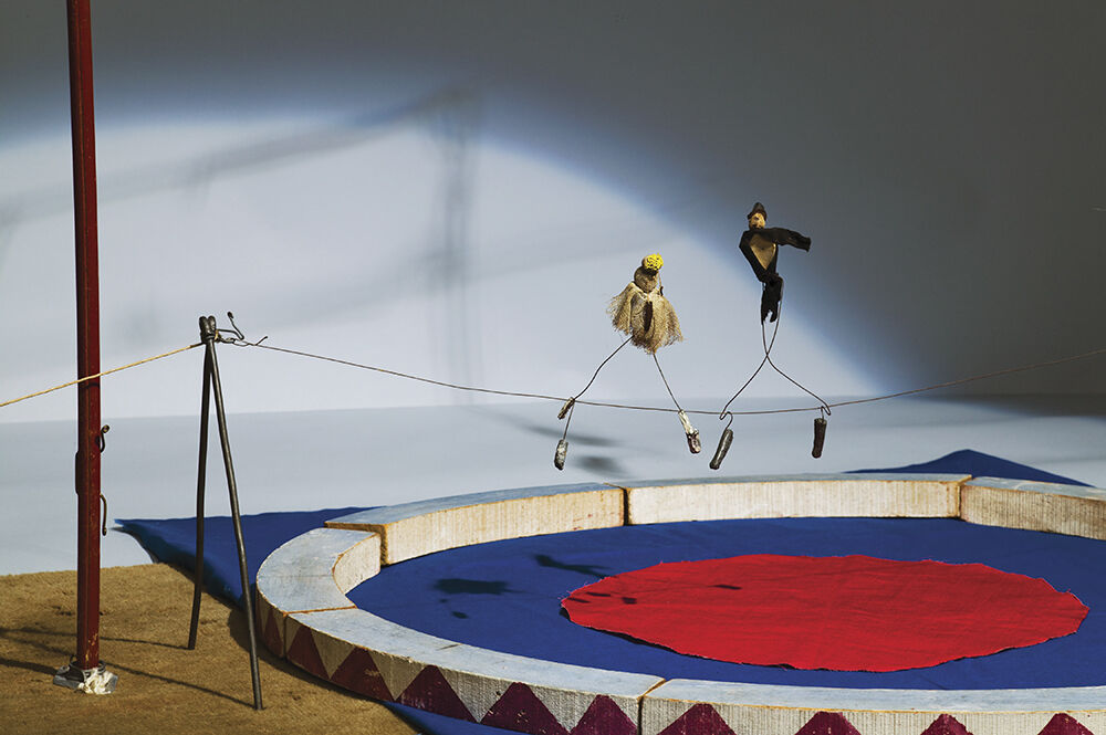 Two small wire puppets dressed as acrobats balance on a tightrope above a miniature circus ring.