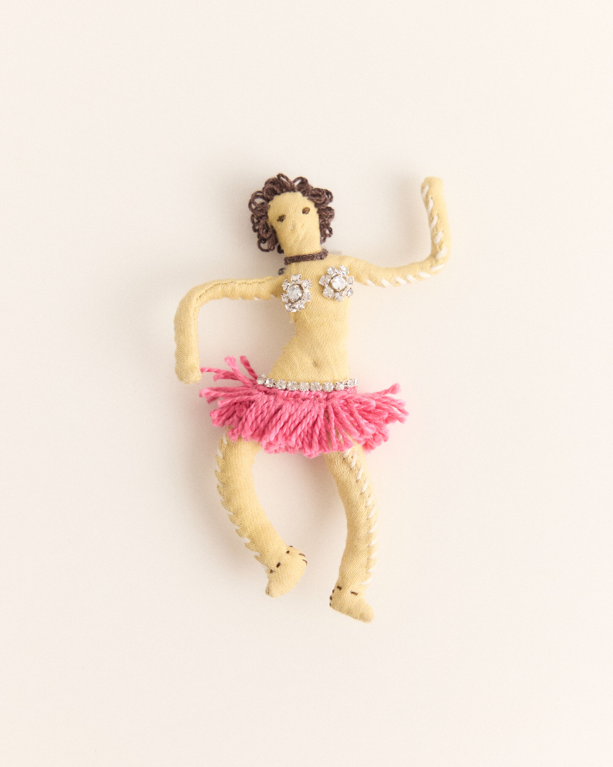 A handmade cloth doll wearing a pink fringe skirt and rhinestone bra, posed dancing.