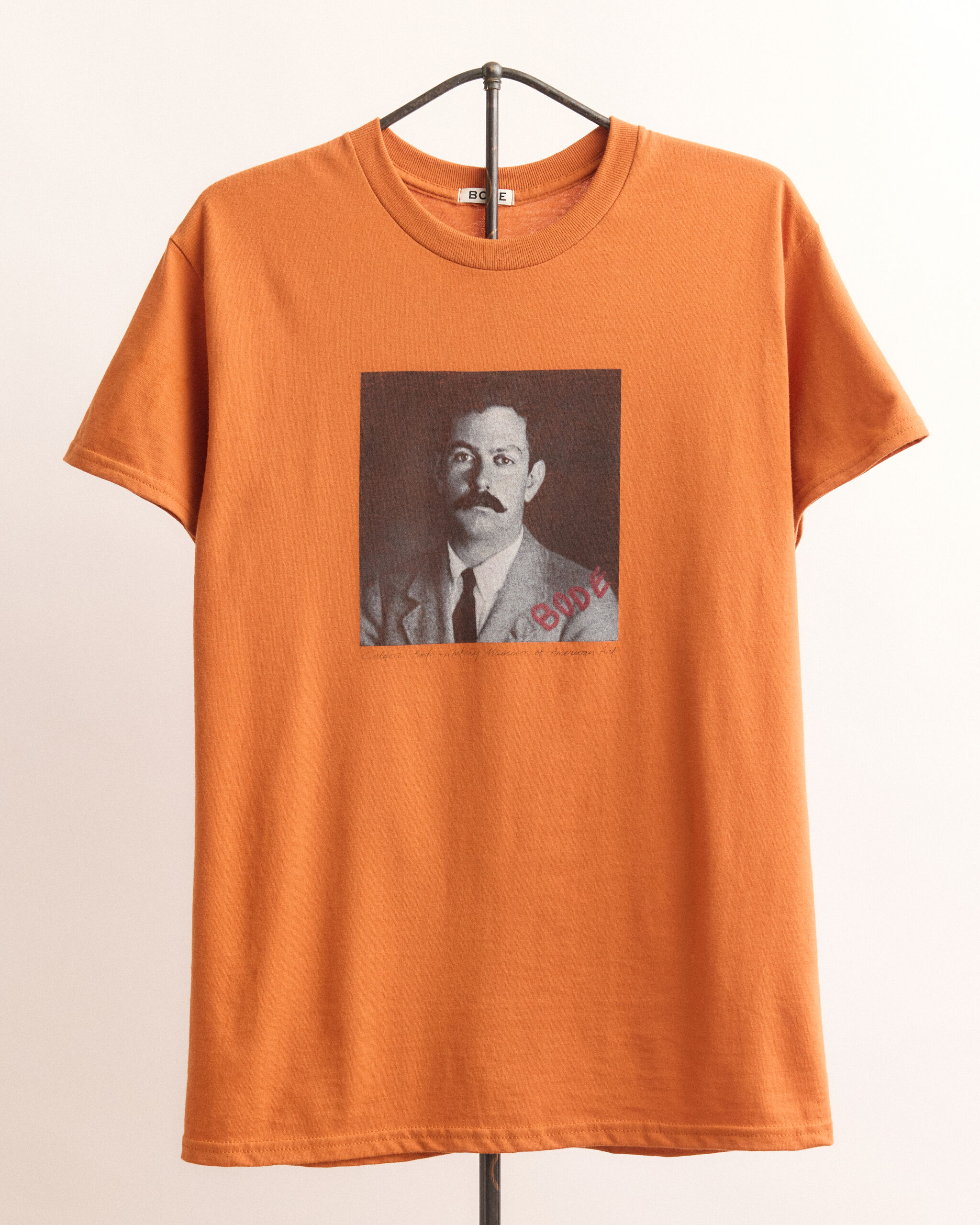 Orange t-shirt hanging on a hanger with a black-and-white portrait and red "BODE" stamp.