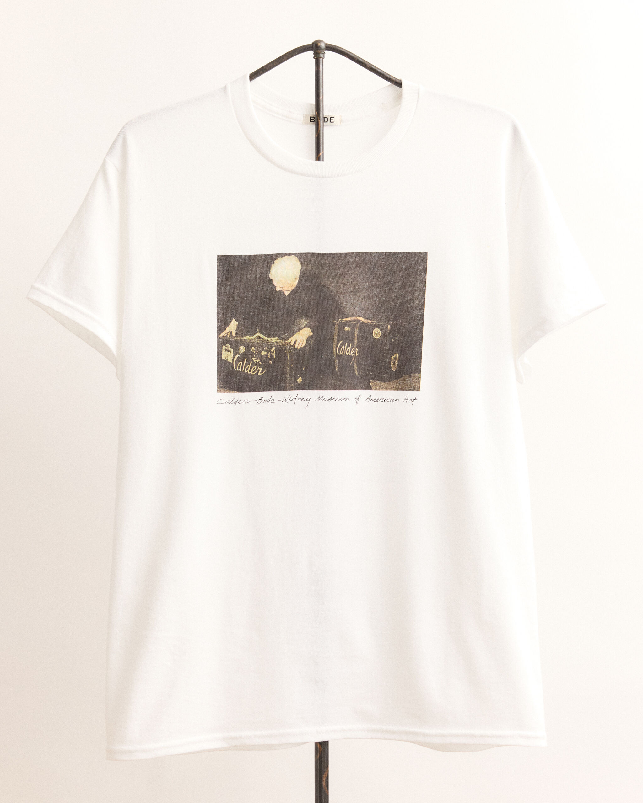 White t-shirt with a printed vintage photo of a person handling suitcases labeled Calder.