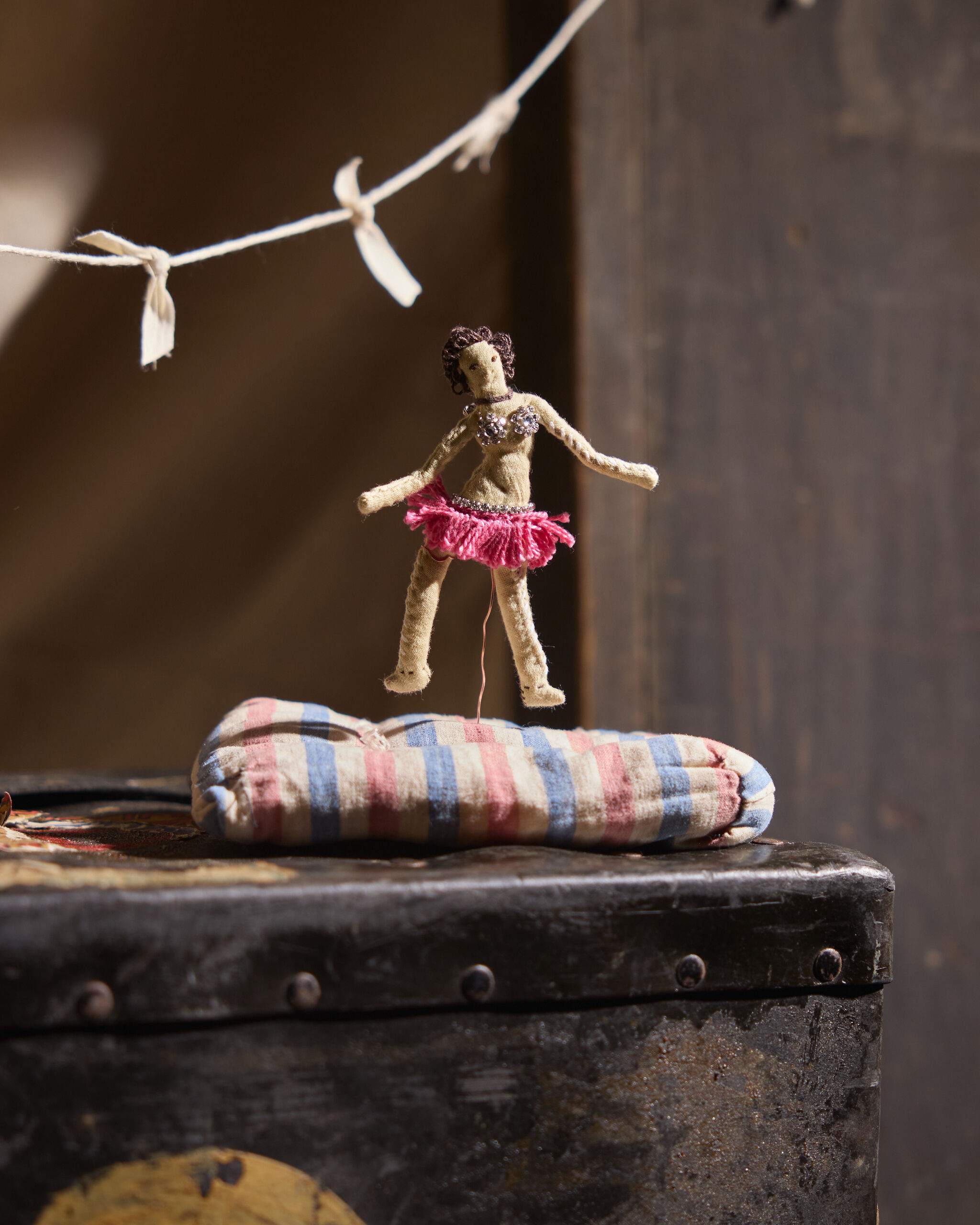A small handmade ballerina doll in a pink skirt hangs above a striped cushion on a worn trunk.