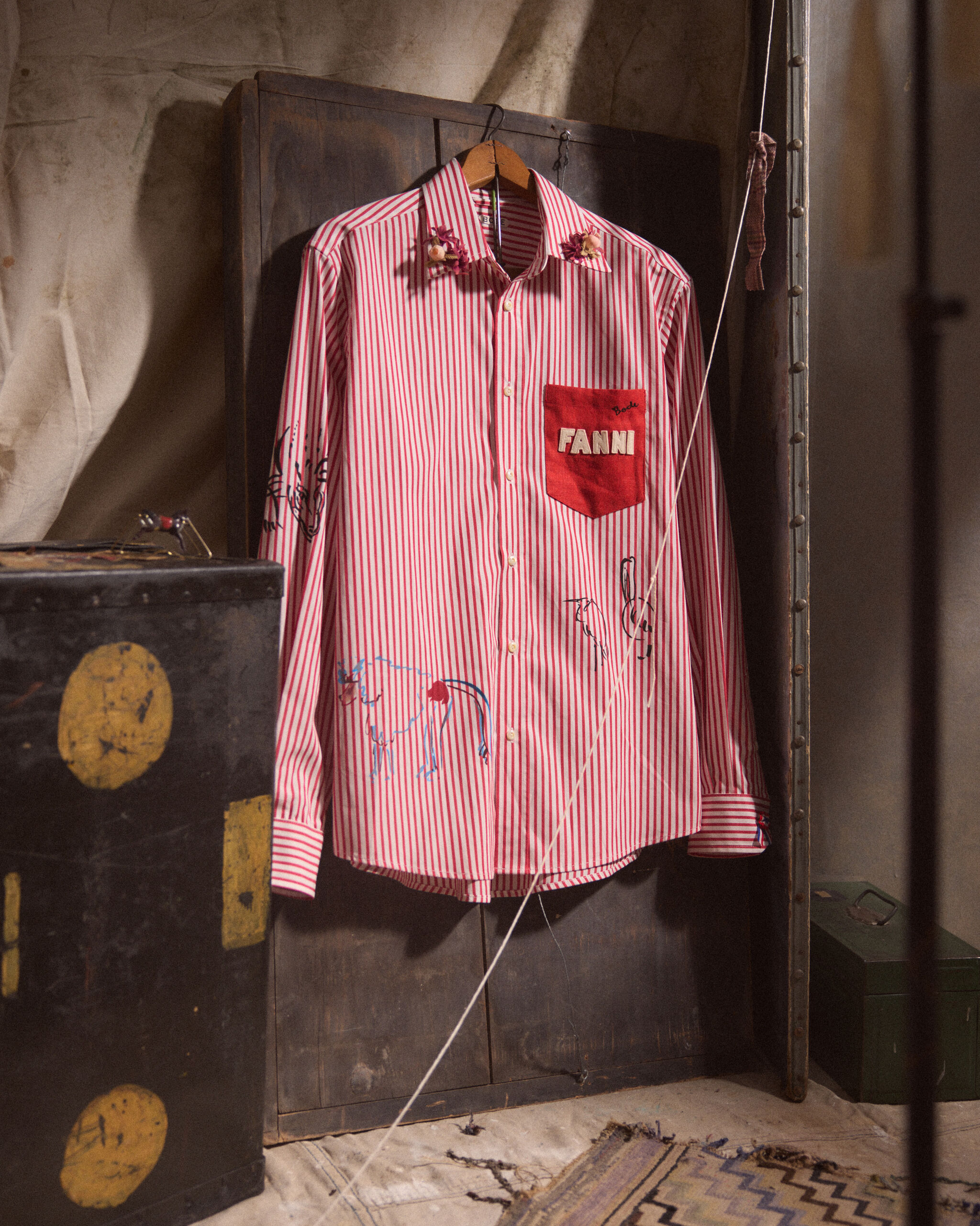 A red-and-white striped shirt hangs on a wooden door, showing a red pocket labeled Fanni and small drawings.