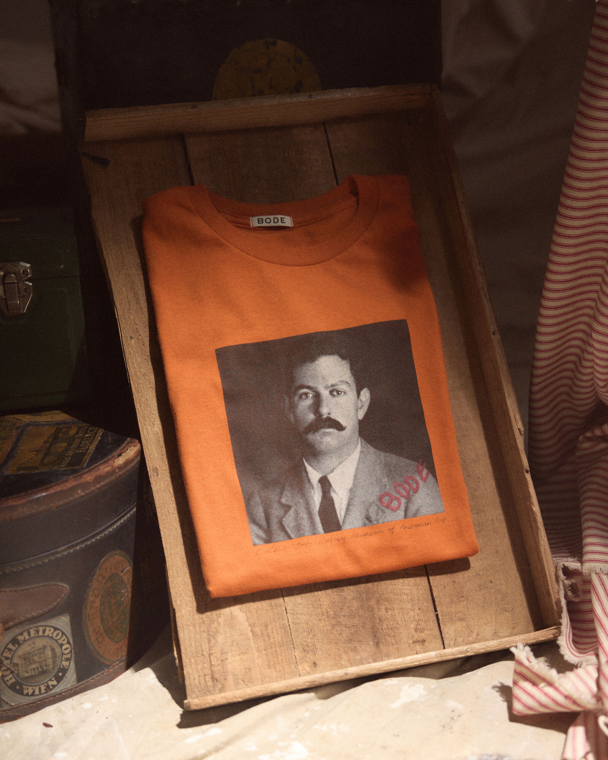 An orange folded T-shirt with a black-and-white portrait and BODE label in a wooden tray.