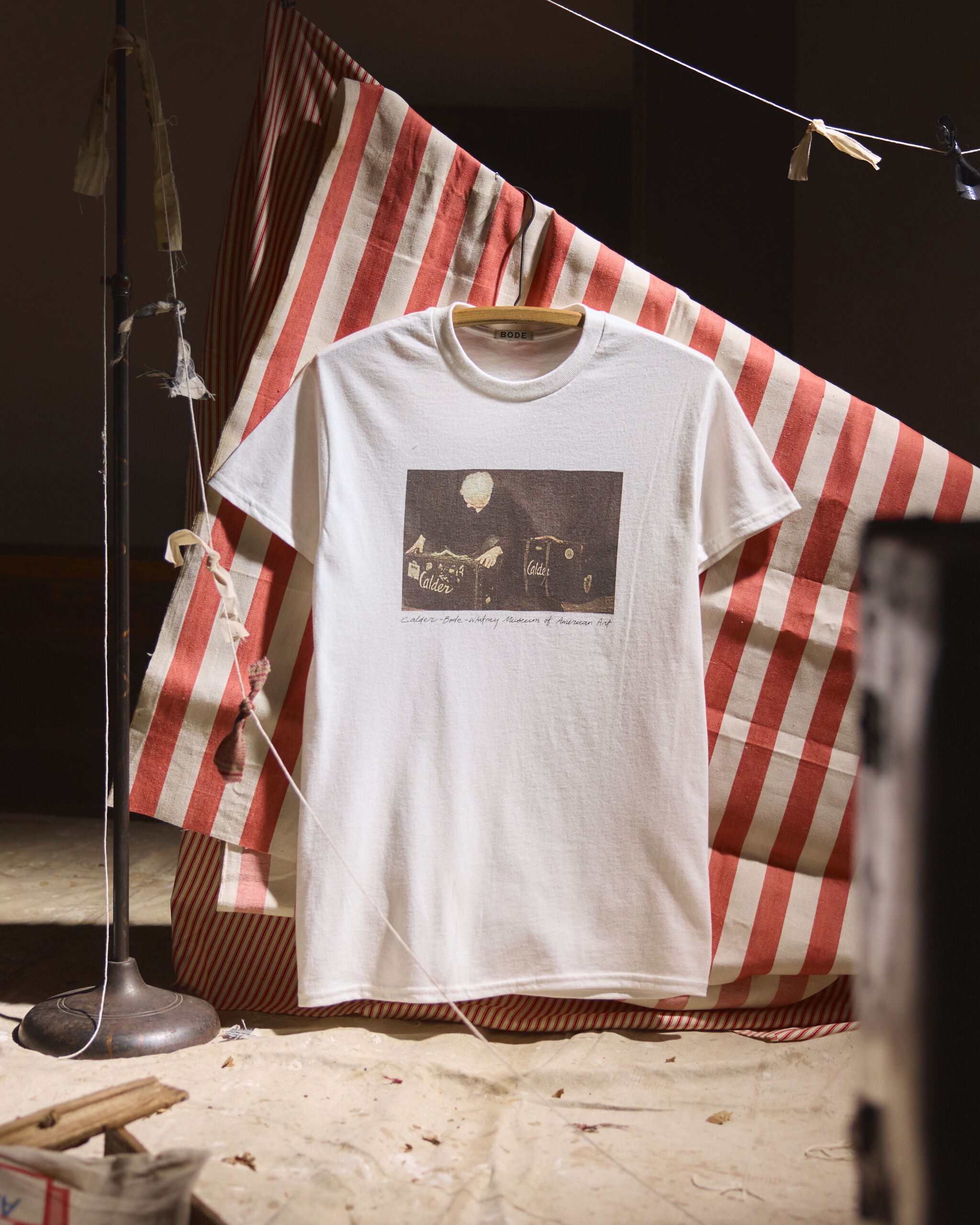 White T-shirt with graphic print hanging on a wooden hanger against red-and-white striped fabric.