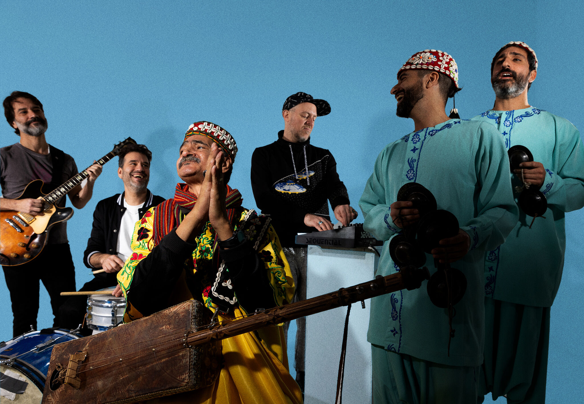 A seated musician in colorful traditional clothes claps while band members play instruments around him.