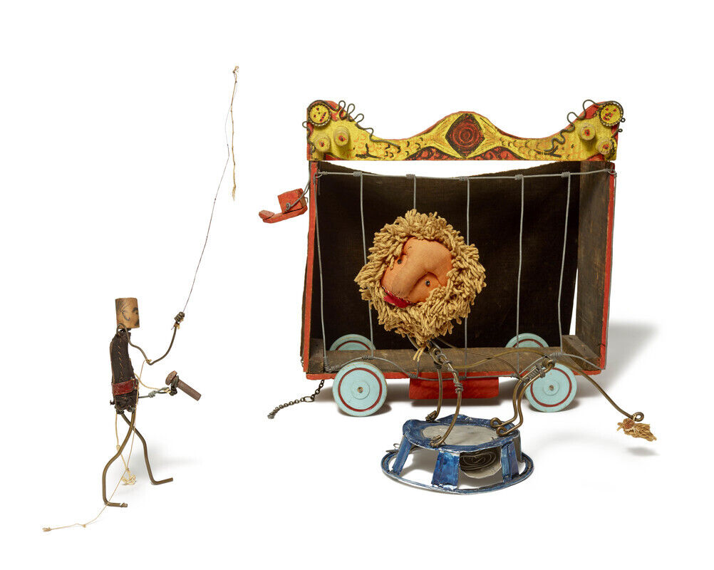 A small wire puppet trainer holds a whip while a stuffed lion hangs inside a colorful wheeled cage.