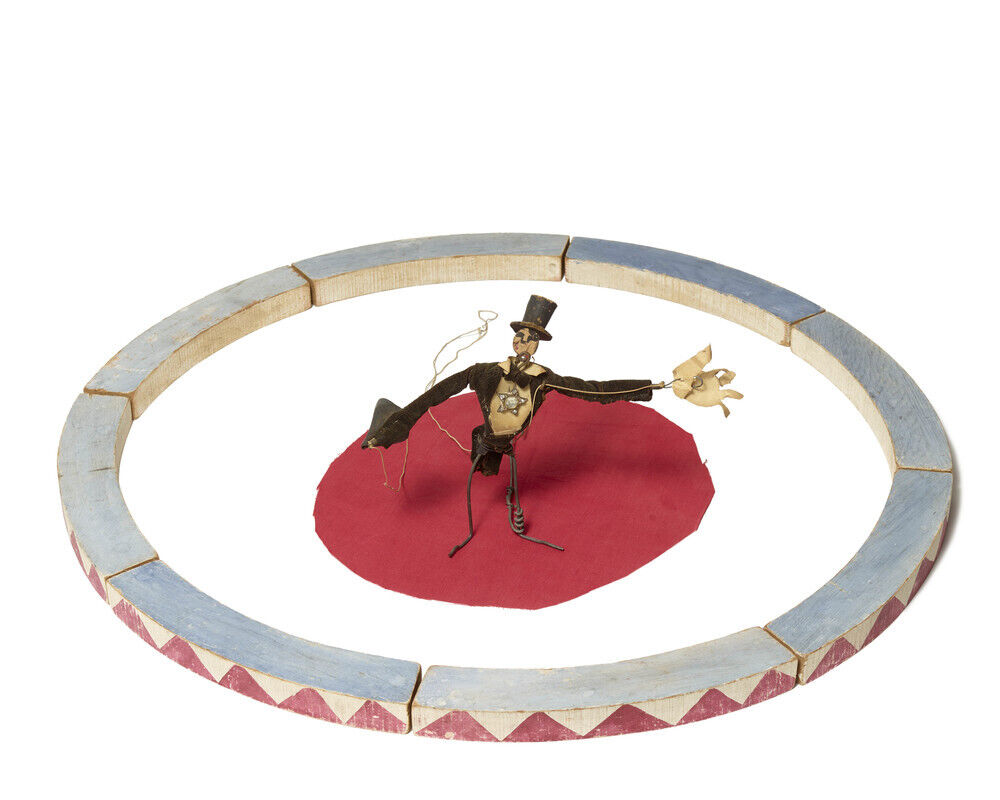 A small toy ringmaster puppet stands in the center of a painted circus ring on a red mat.