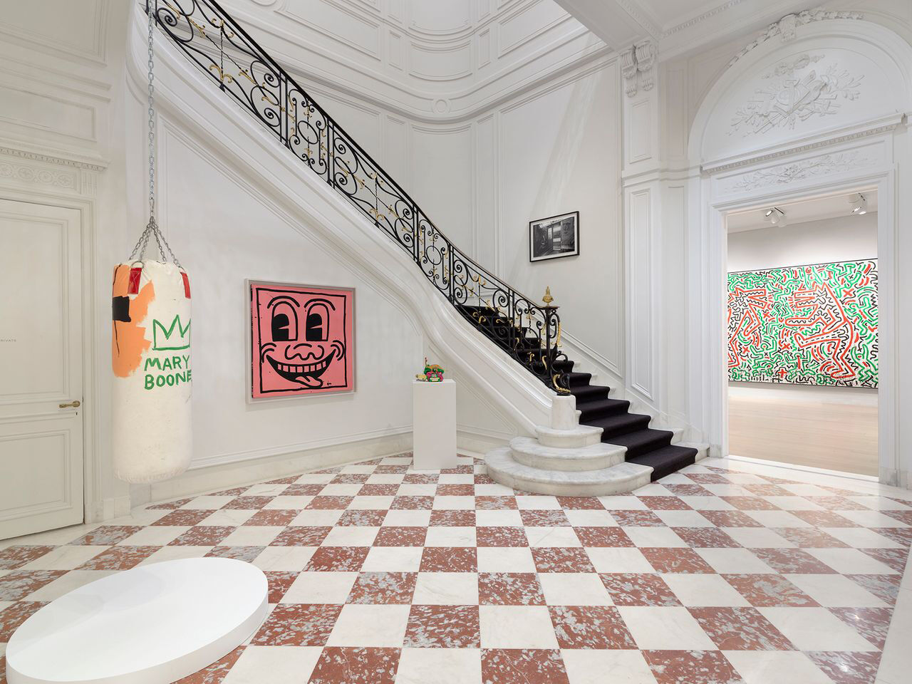 Five art pieces are displayed inside of a historic townhouse with a marble staircase, decorative arches, and tall ceilings.