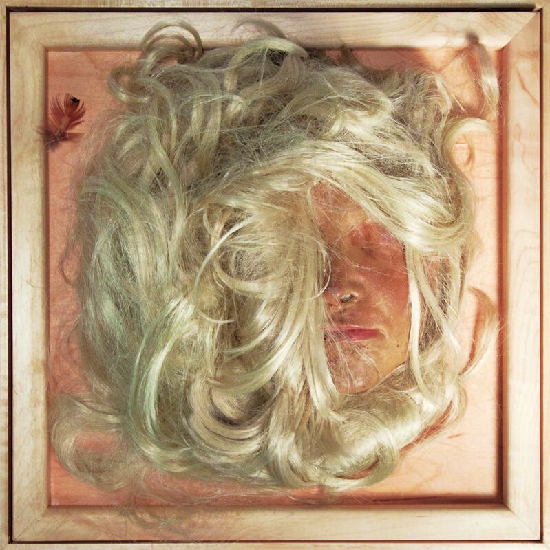 A blonde wig partially covering a human-like face mask mounted inside a shallow wooden frame.