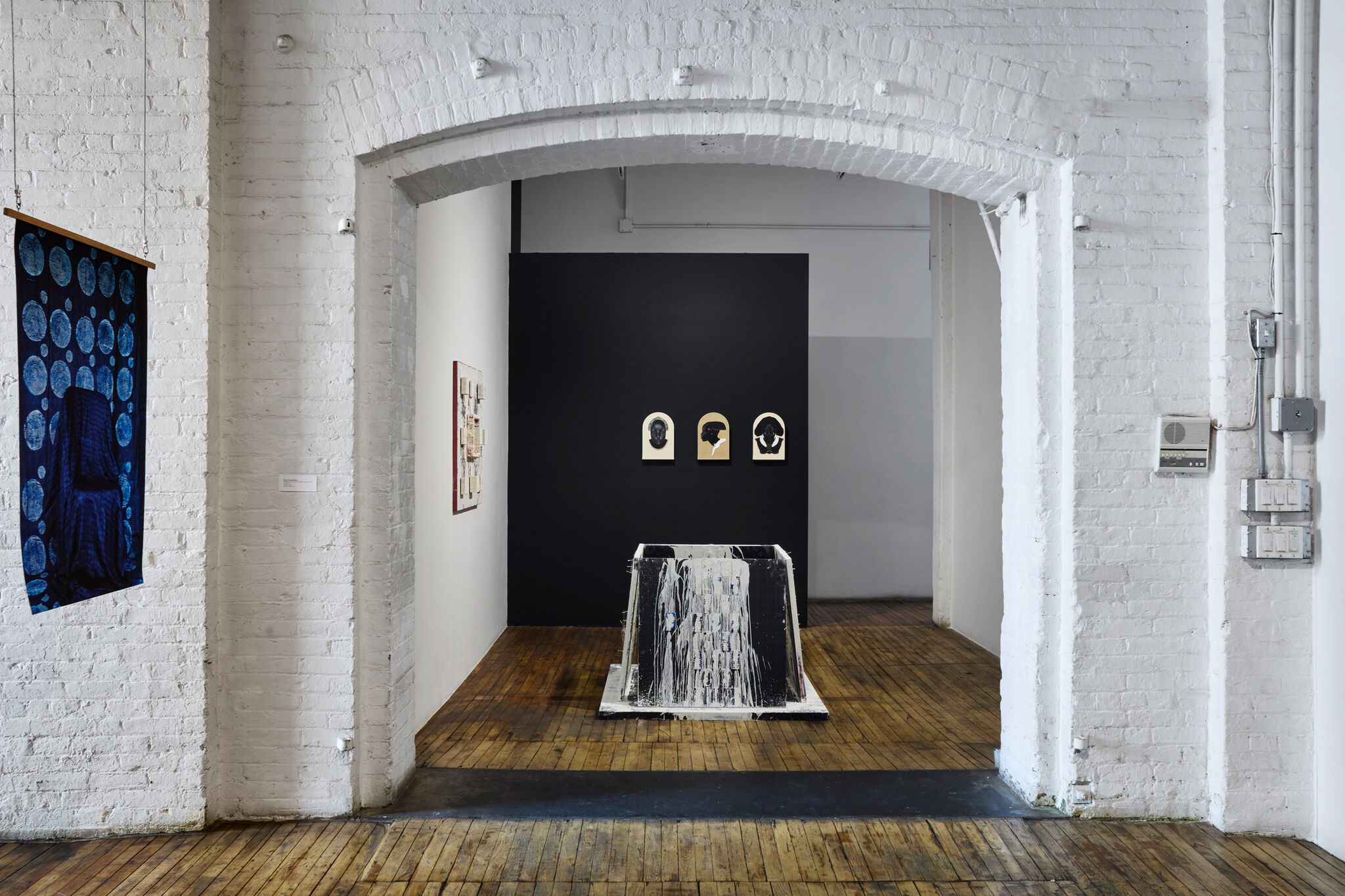 Central paint-splattered sculpture sits on wooden floor beneath three small framed silhouette portraits on dark wall.