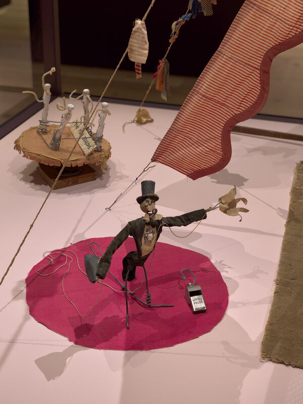 A wire and fabric marionette in a top hat balances on one leg on a pink circle.