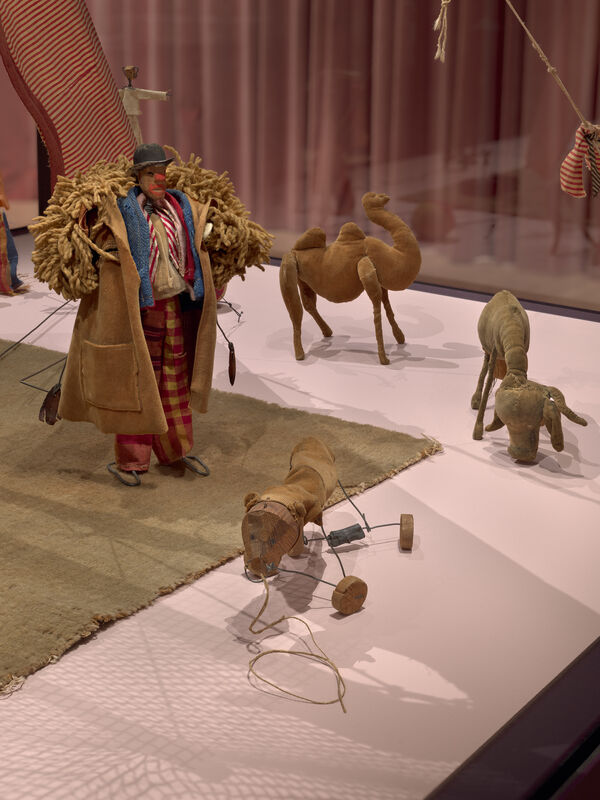 A cloth puppet man pulls a wheeled toy animal while stuffed camels stand nearby.