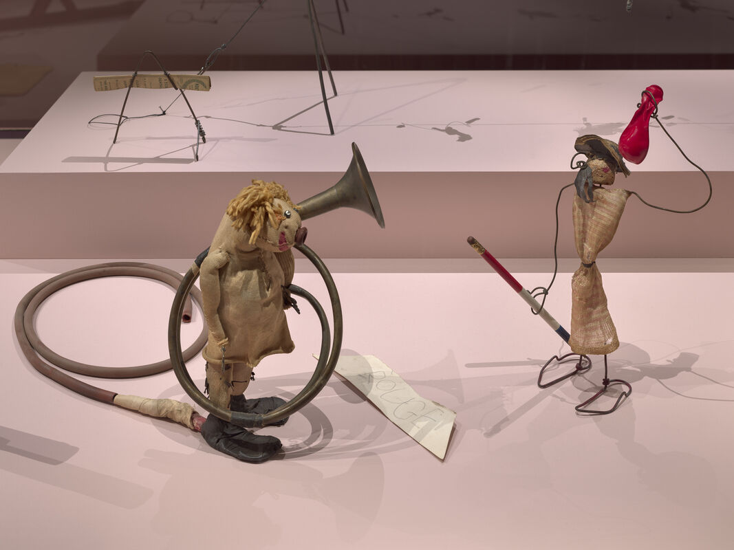 Two handmade puppet figures made from fabric and wire interact, one with a horn and coiled hose.