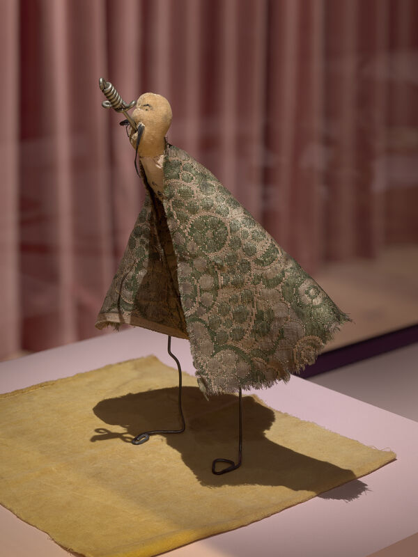 Small handmade figure with a patterned green cloak, wire legs, and a cork head with spiral nose.