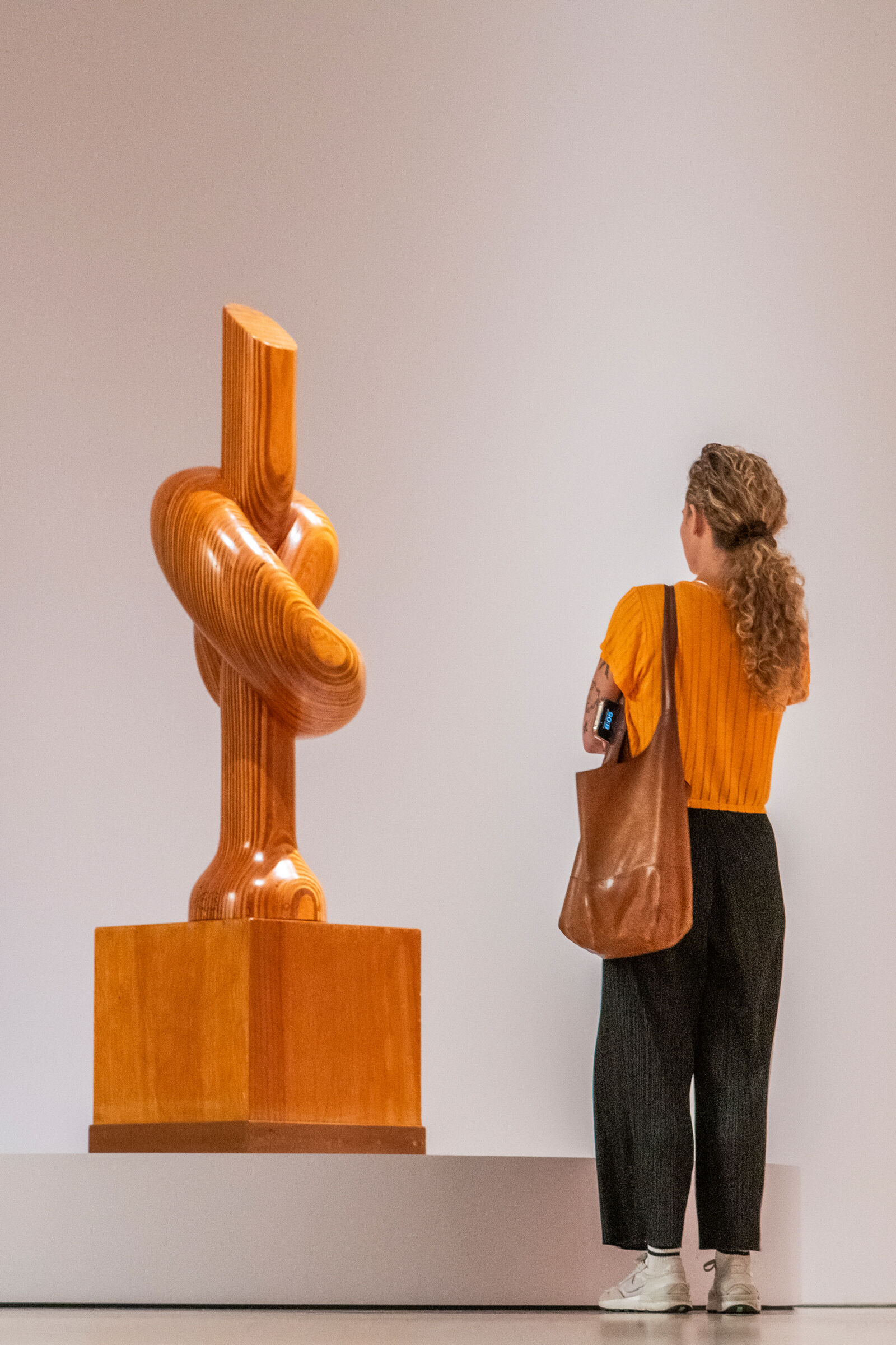 A woman stands to the right of a wooden sculpture observing it. The sculpture is tall, light brown and shaped like a knot. It stands on a white round platform against one wall in the Sixties Surreal gallery.