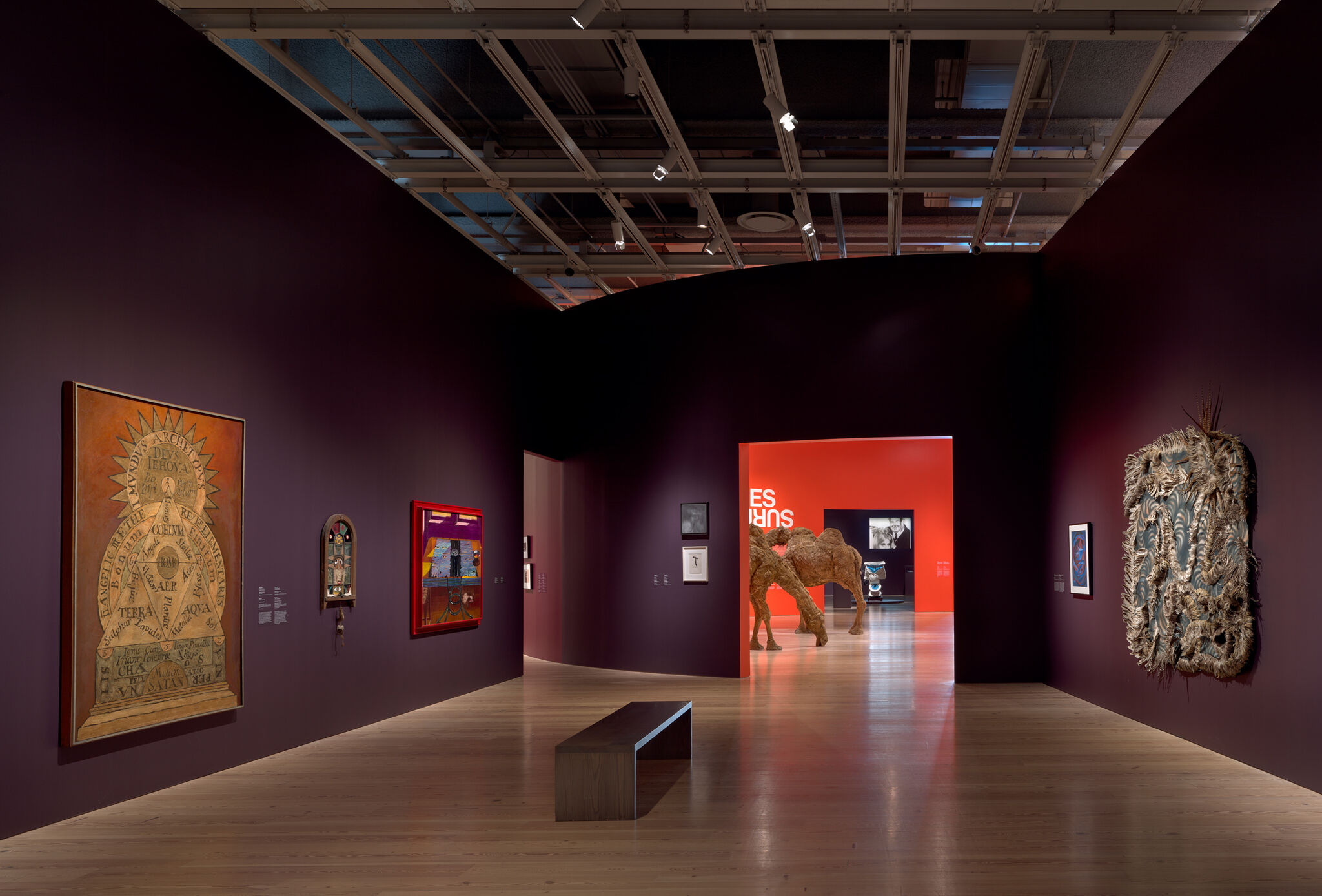 Gallery with dark purple walls displaying framed artworks and a large camel sculpture in an adjacent room.