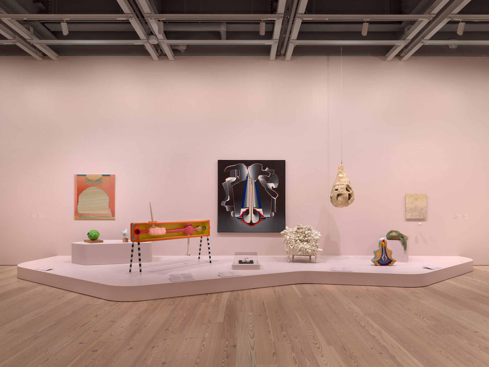A gallery display of sculptures and paintings arranged on a low platform against a pink wall.