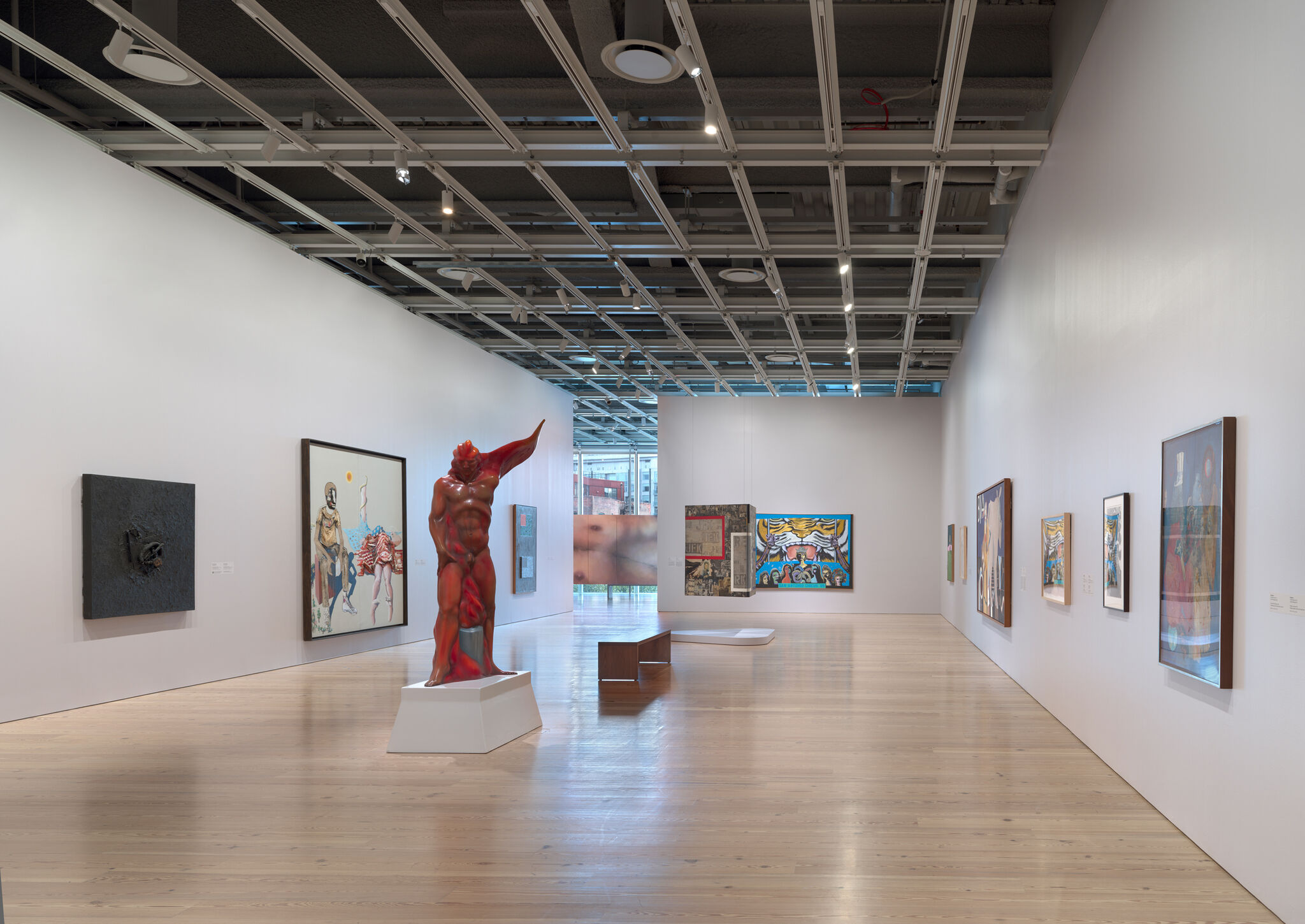 A red muscular sculpture stands on a white pedestal in a spacious gallery surrounded by paintings.