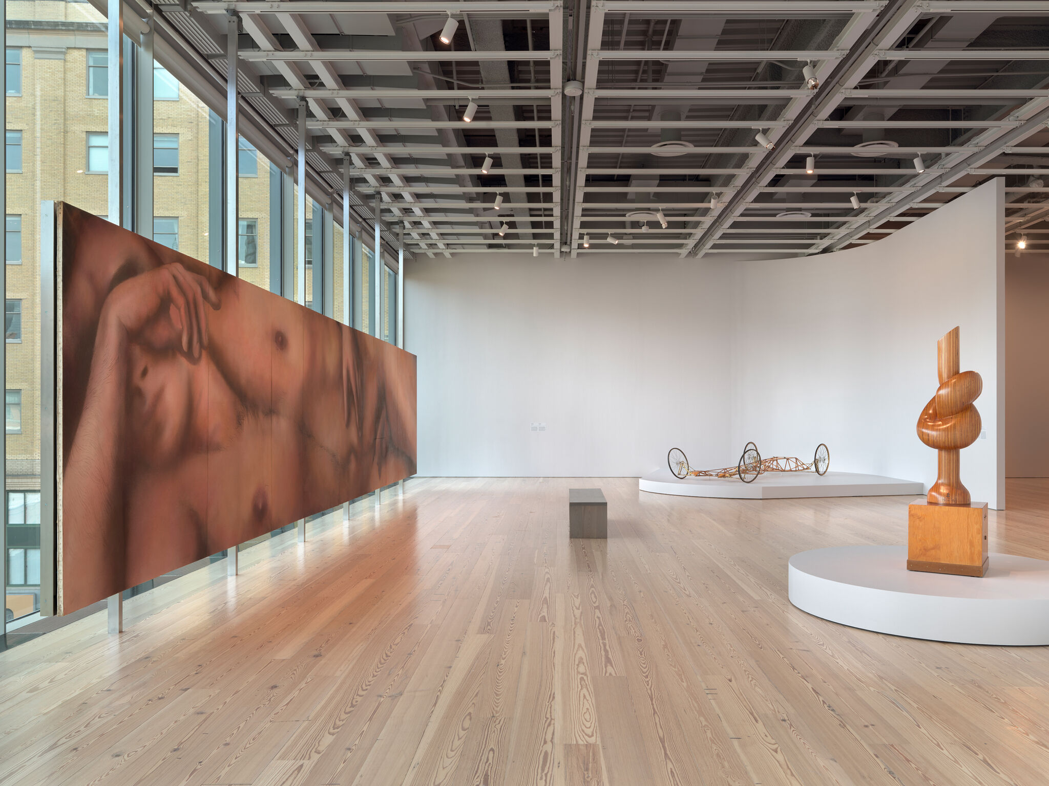 A spacious gallery features a long reclining nude painting along windows and a wooden abstract sculpture.