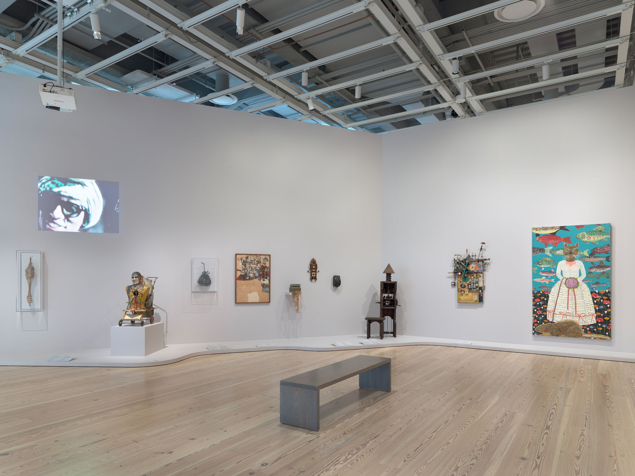 A bright gallery room displaying diverse sculptures, paintings, and a projected video on a white wall.
