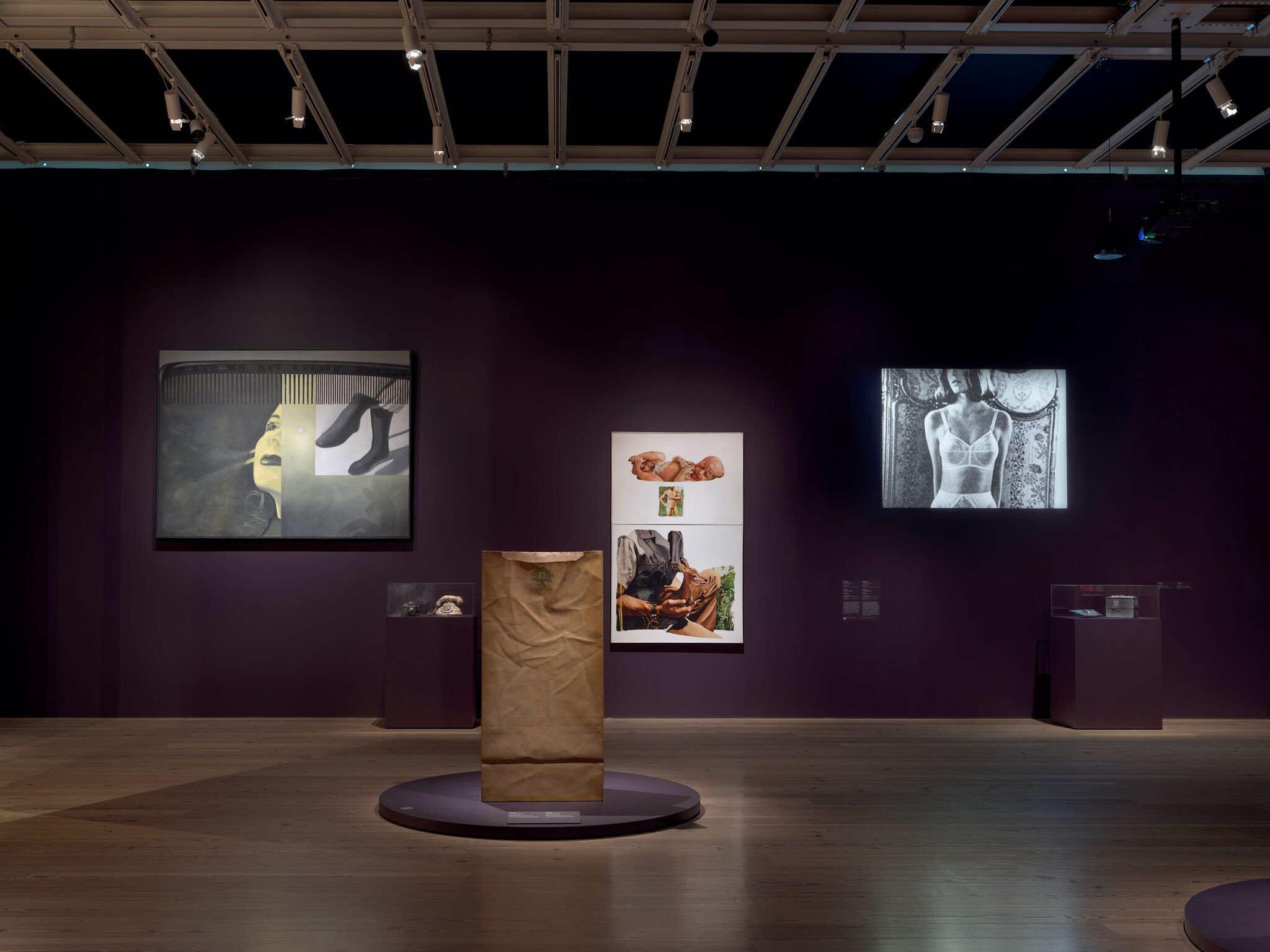 A tall brown paper bag sculpture stands on a round pedestal in a dim gallery with framed artworks.