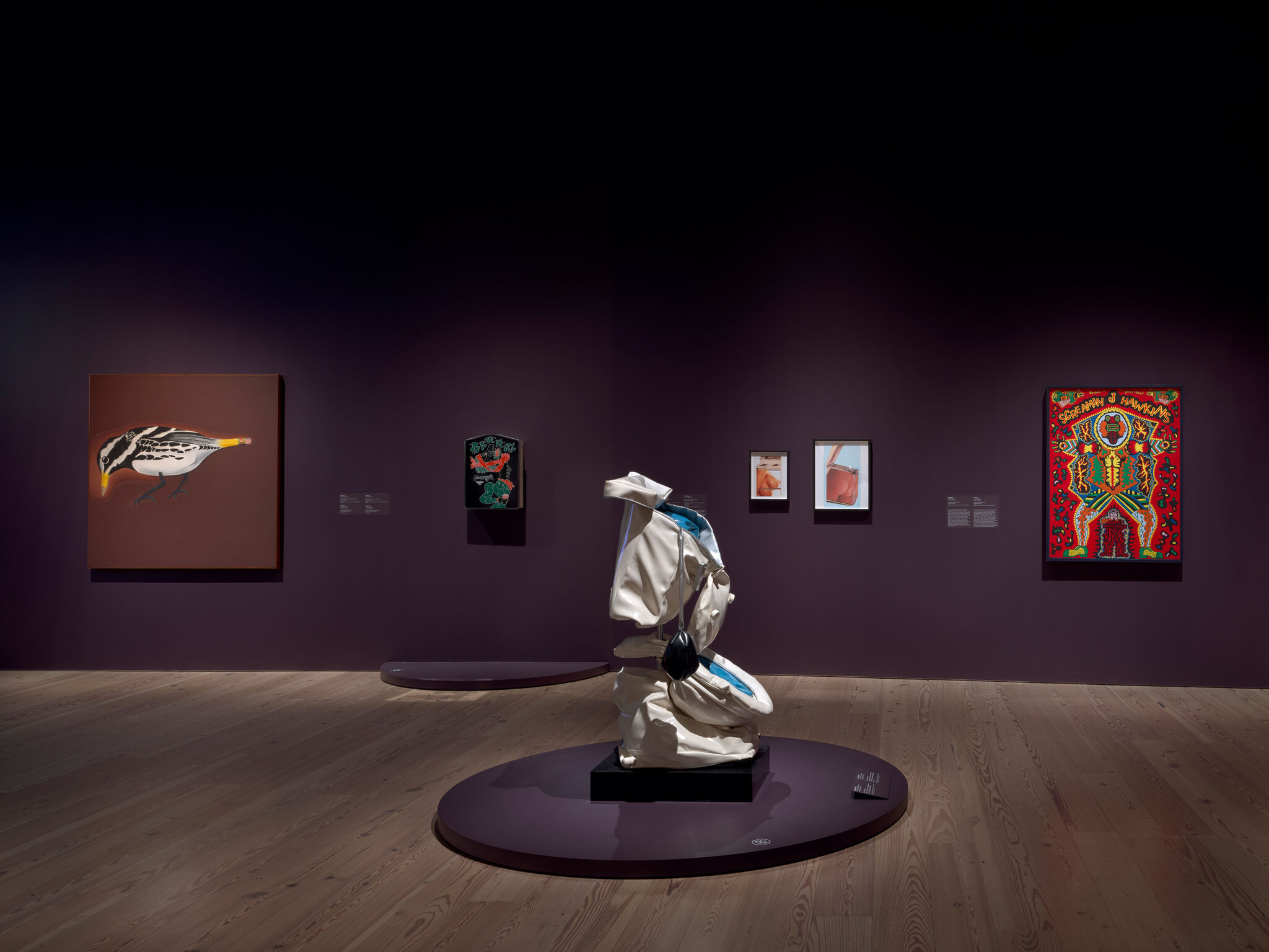 A crushed toilet sculpture stands on a round platform surrounded by colorful paintings on purple walls.