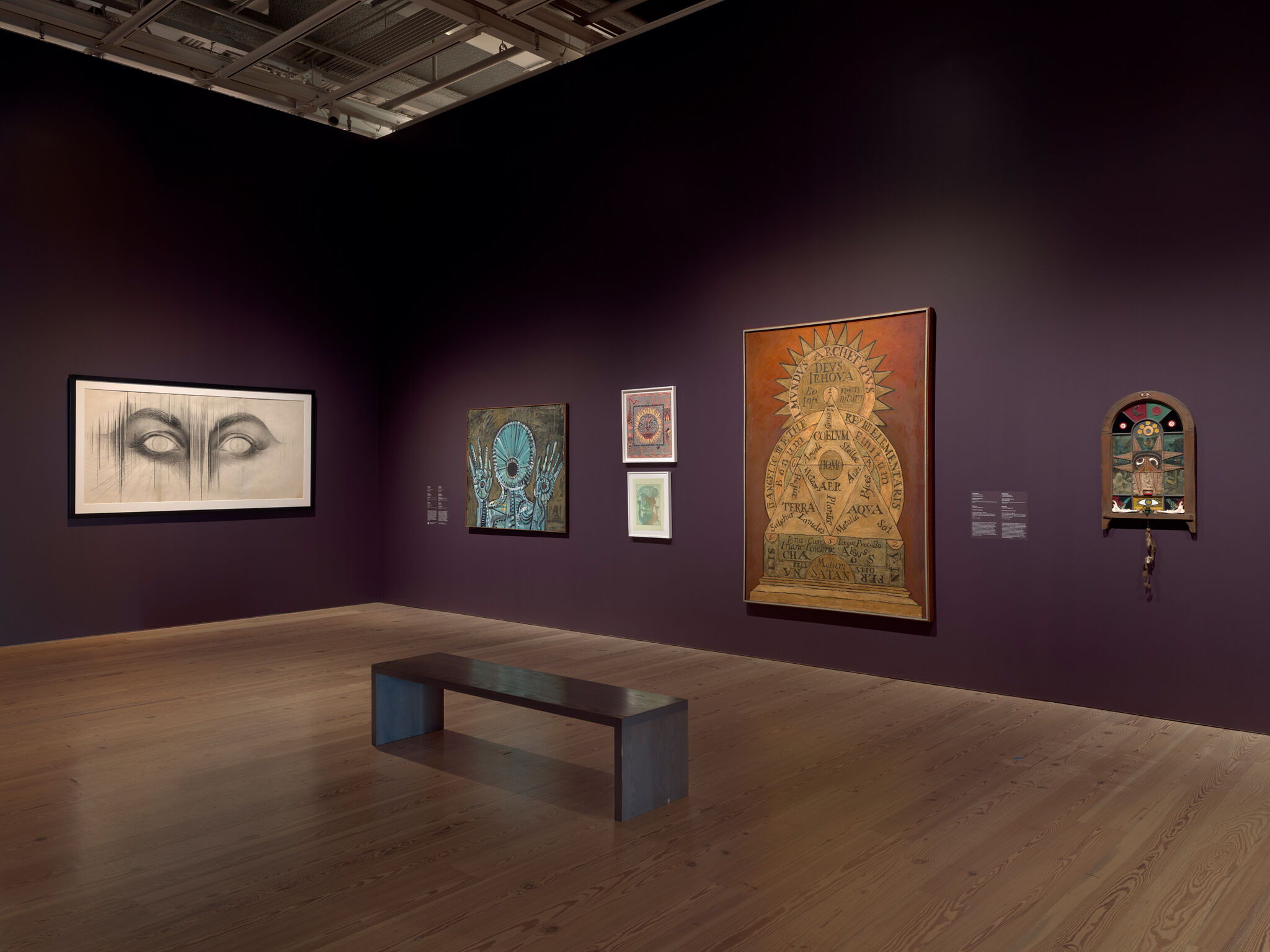 Gallery room with several framed artworks on a deep purple wall and a wooden bench.