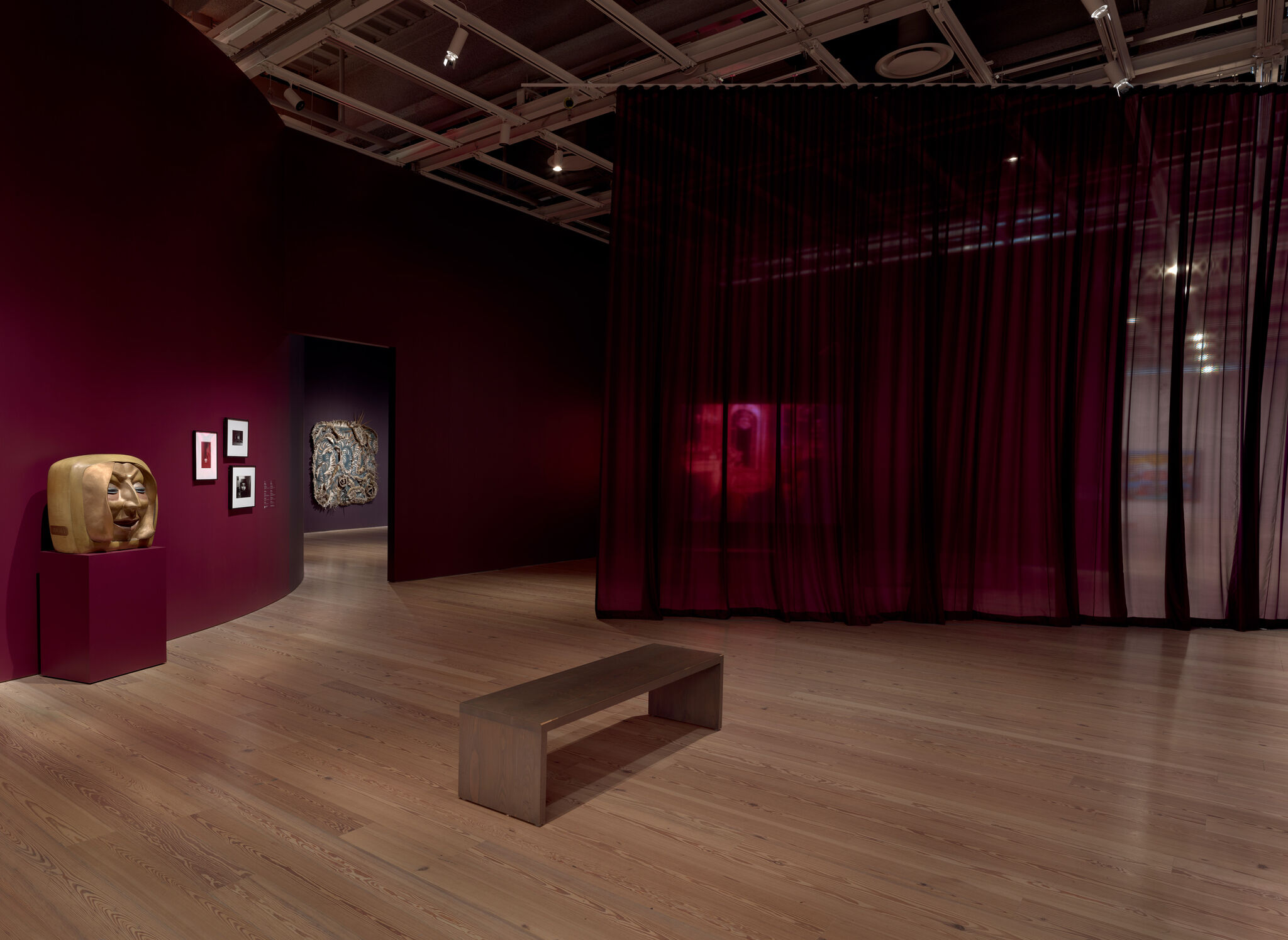 A wooden bench faces a dim gallery with a gold face sculpture and sheer burgundy curtain.