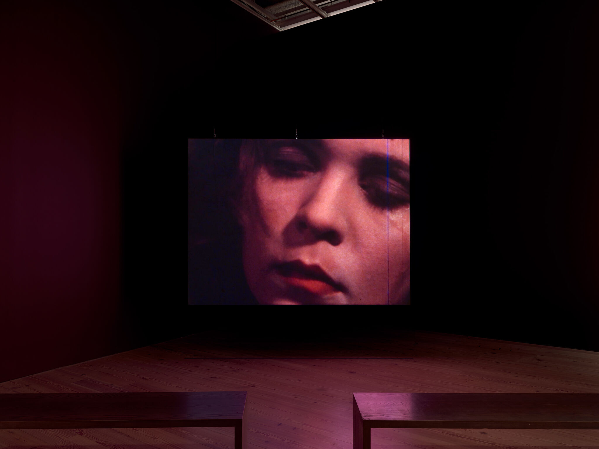 Large projected close-up of a woman's face displayed on a dark gallery wall with empty benches.