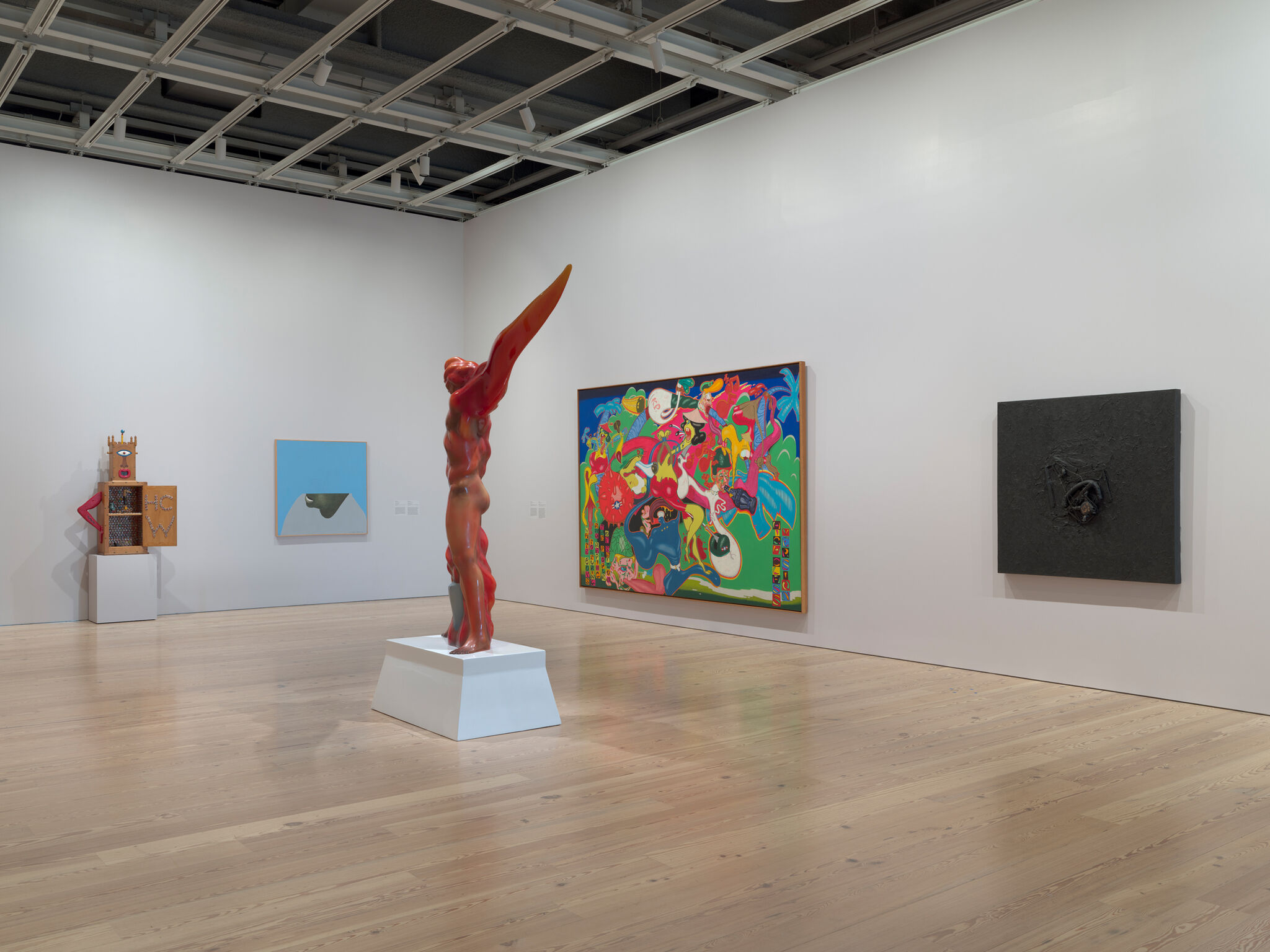 Red abstract human-like sculpture with extended arm stands on a white pedestal in a gallery.