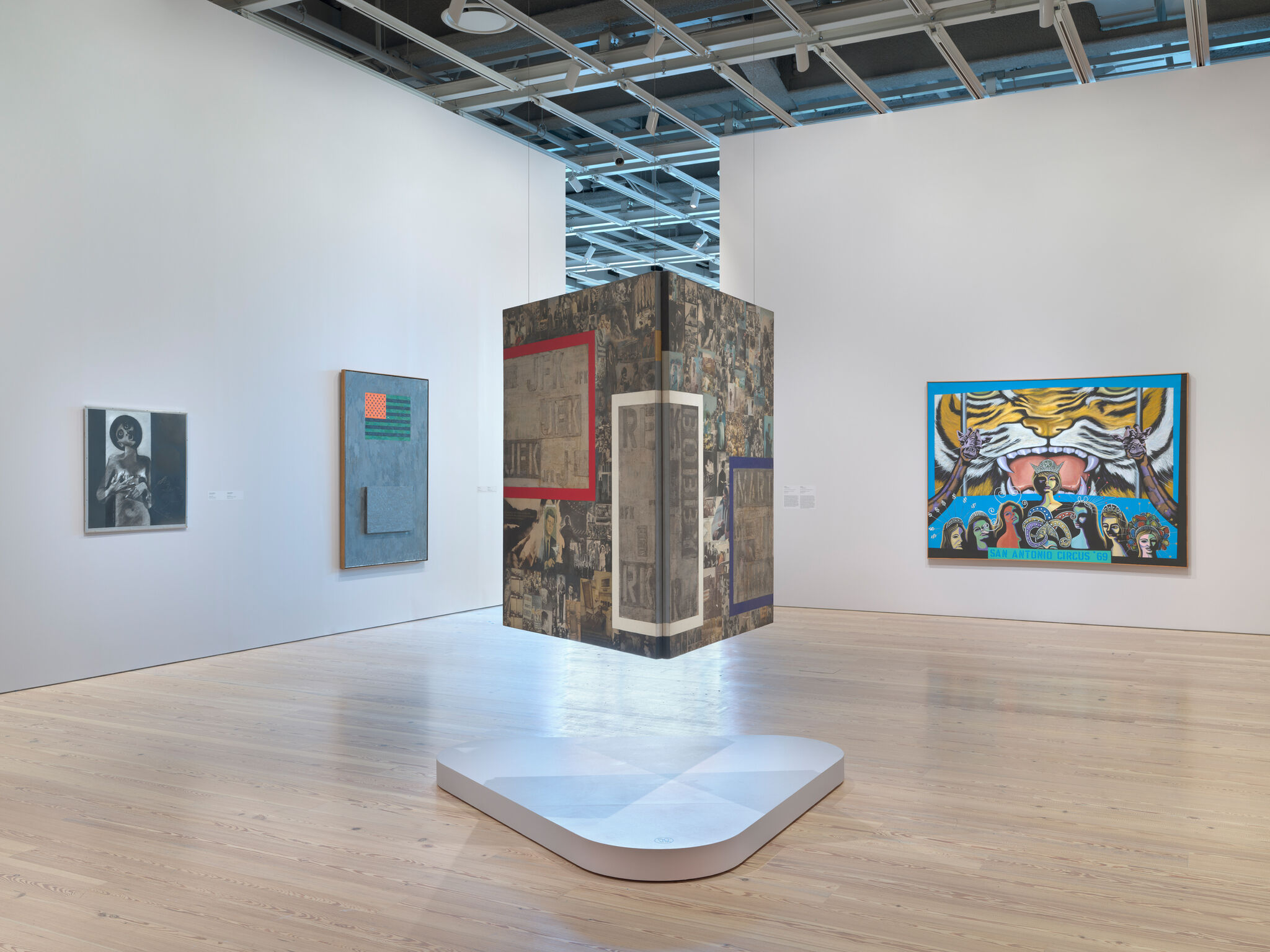 A suspended cube installation hangs above a white pedestal in a gallery.