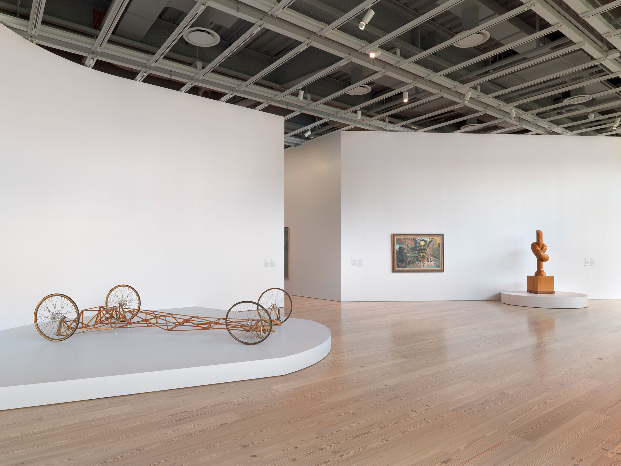 A gallery displaying a wooden lattice-wheeled sculpture, framed painting, and wooden abstract sculpture.