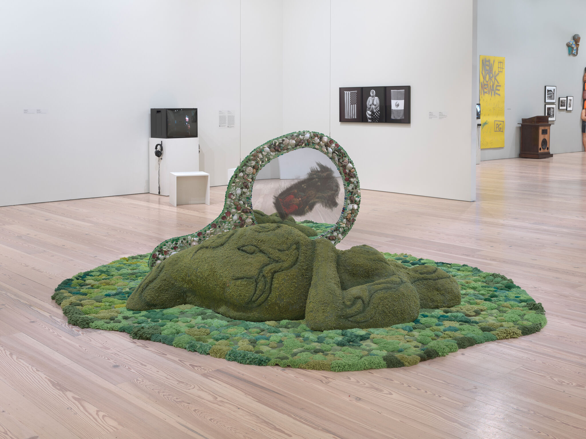 A large moss-covered reclining sculpture on a round green mound with a reflective circular frame.