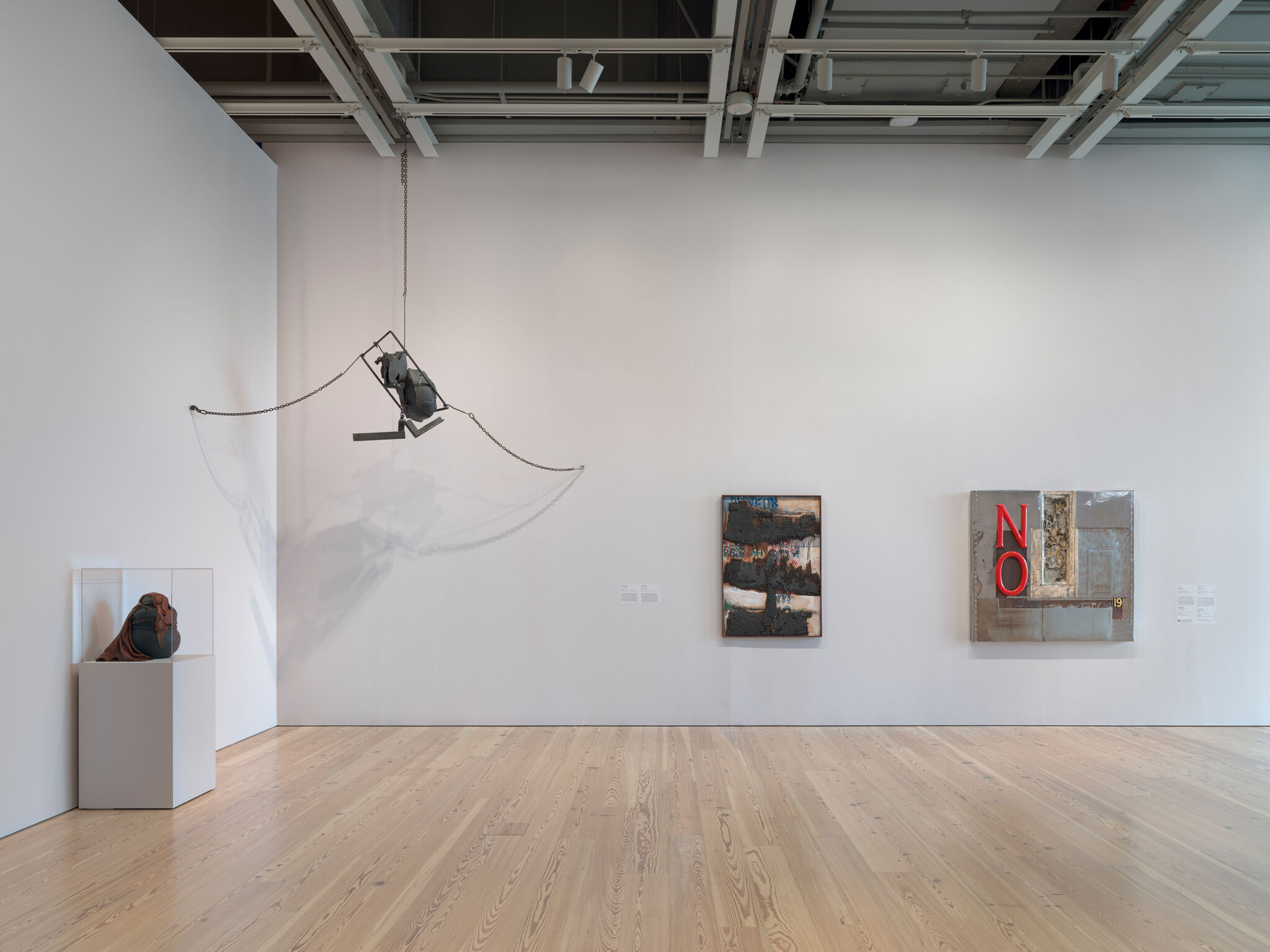 A museum gallery wall with a hanging metal sculpture and artworks, including a piece showing red letters "NO".