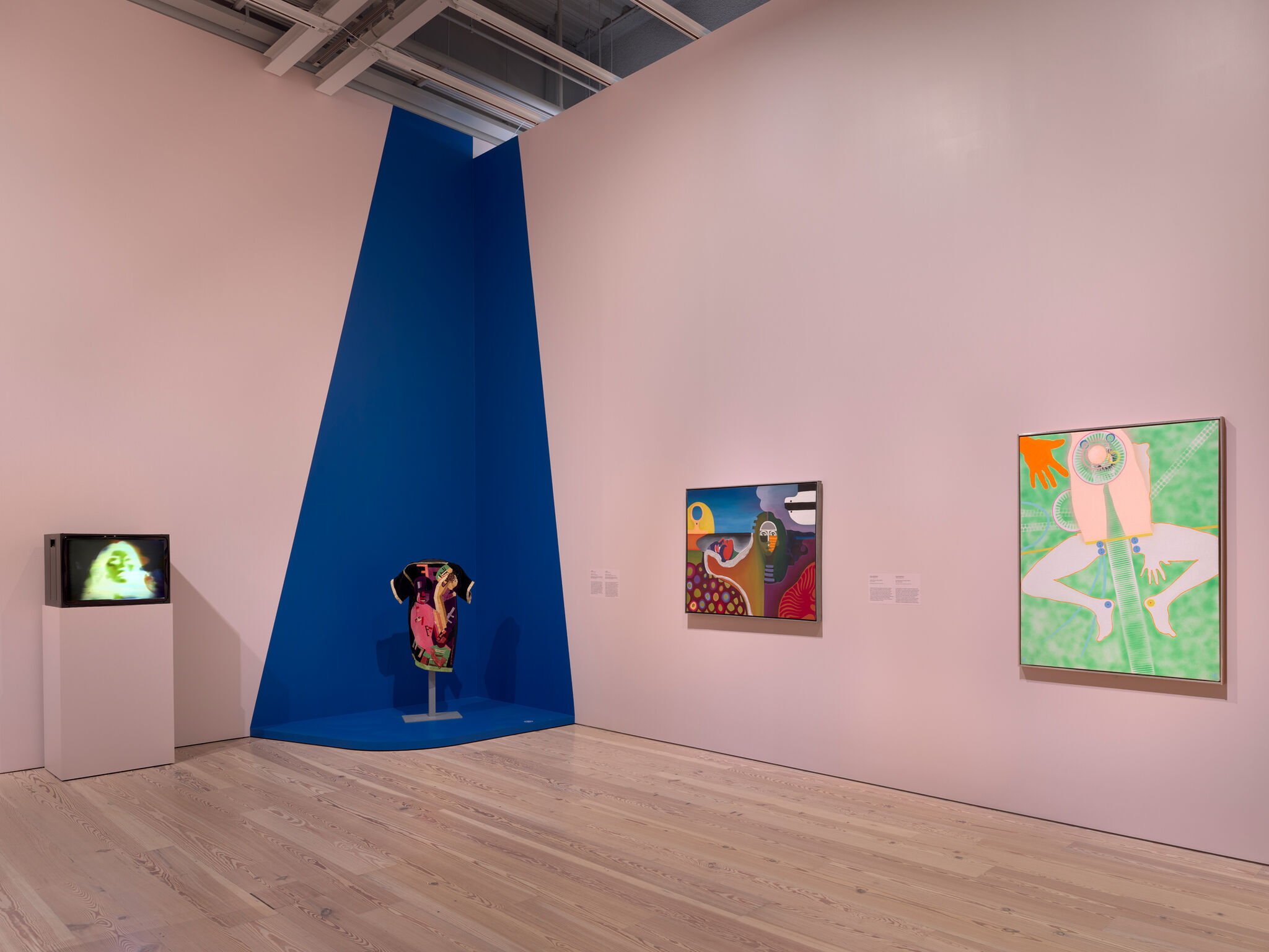 A pink gallery room with colorful paintings, a video monitor, and a blue corner displaying a sculpture on a pedestal.