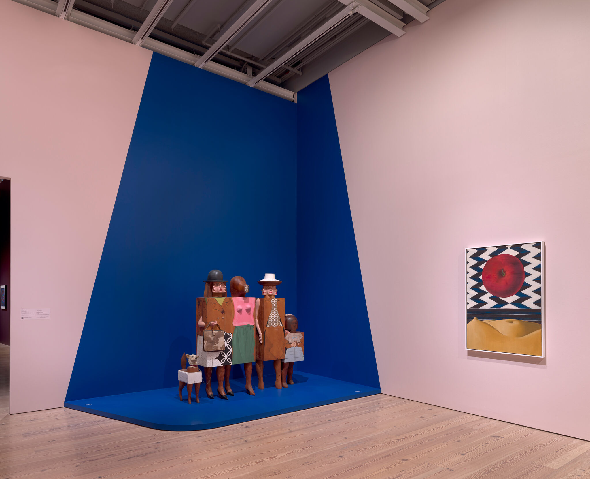 A group of stylized wooden figures stands on a blue triangular platform against gallery walls.