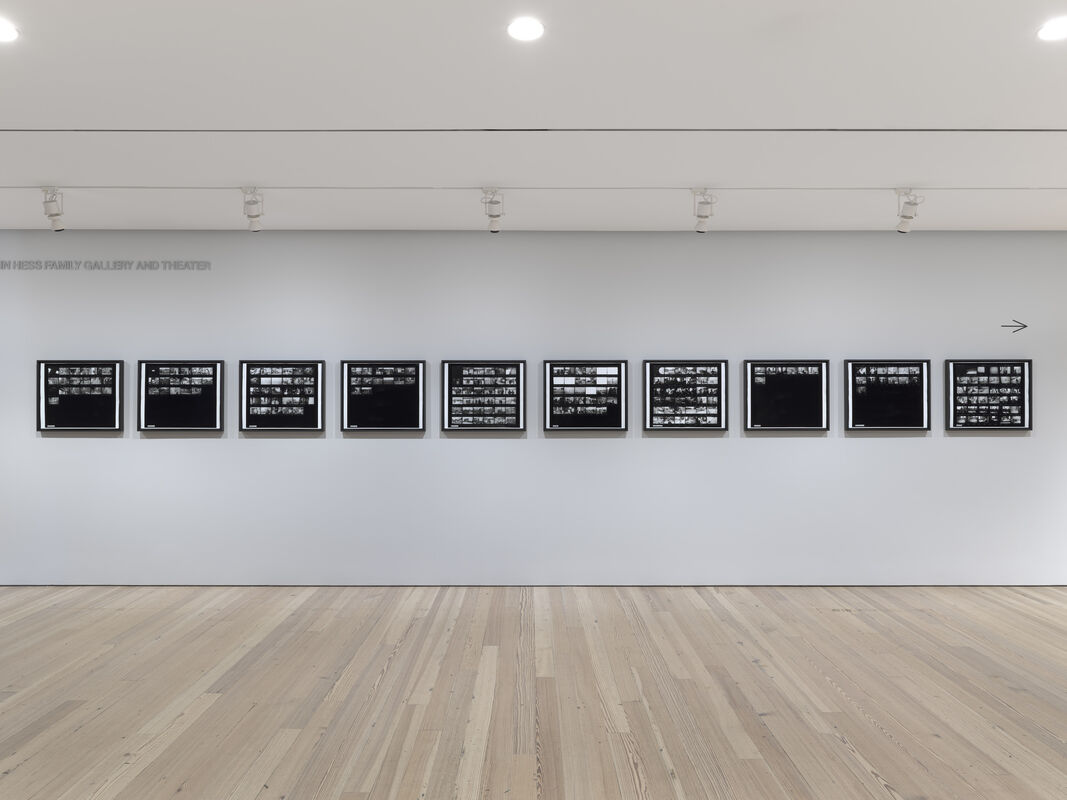 A gallery wall displays eleven framed black-and-white contact sheet photographs evenly spaced above a wooden floor.