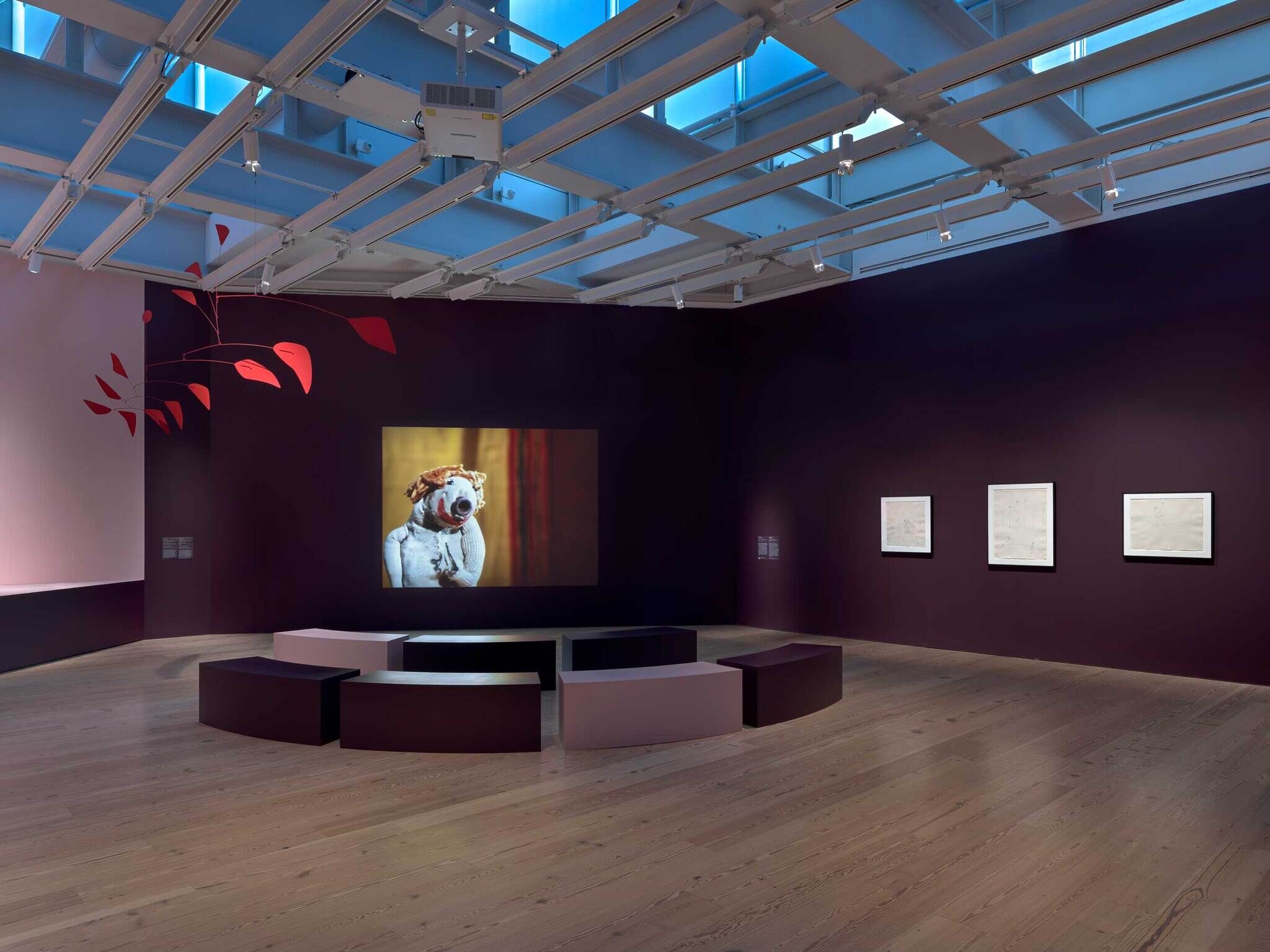 Dark gallery room with circular benches facing a projected puppet figure on a wall screen.