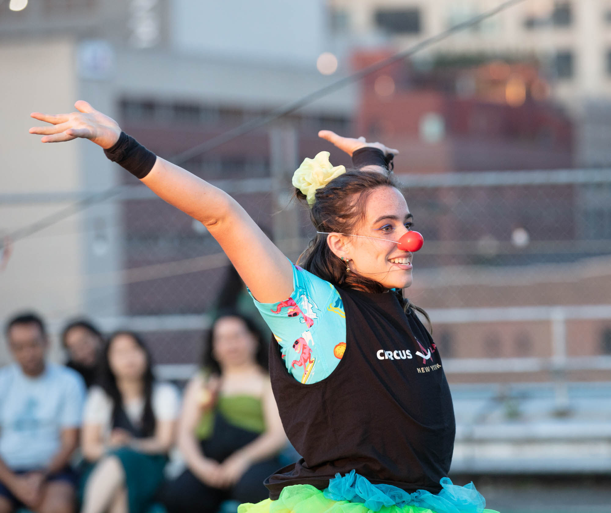 A performer from the Circus Academy of New York smiling with arms outstretched, wearing a red clown nose, colorful costume, and green tulle skirt. The performer is outdoors with a blurred audience and city buildings in the background.