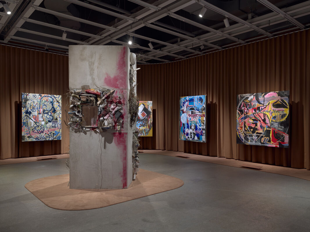 A gallery room with a central concrete column covered in mixed objects and colorful abstract paintings on walls.