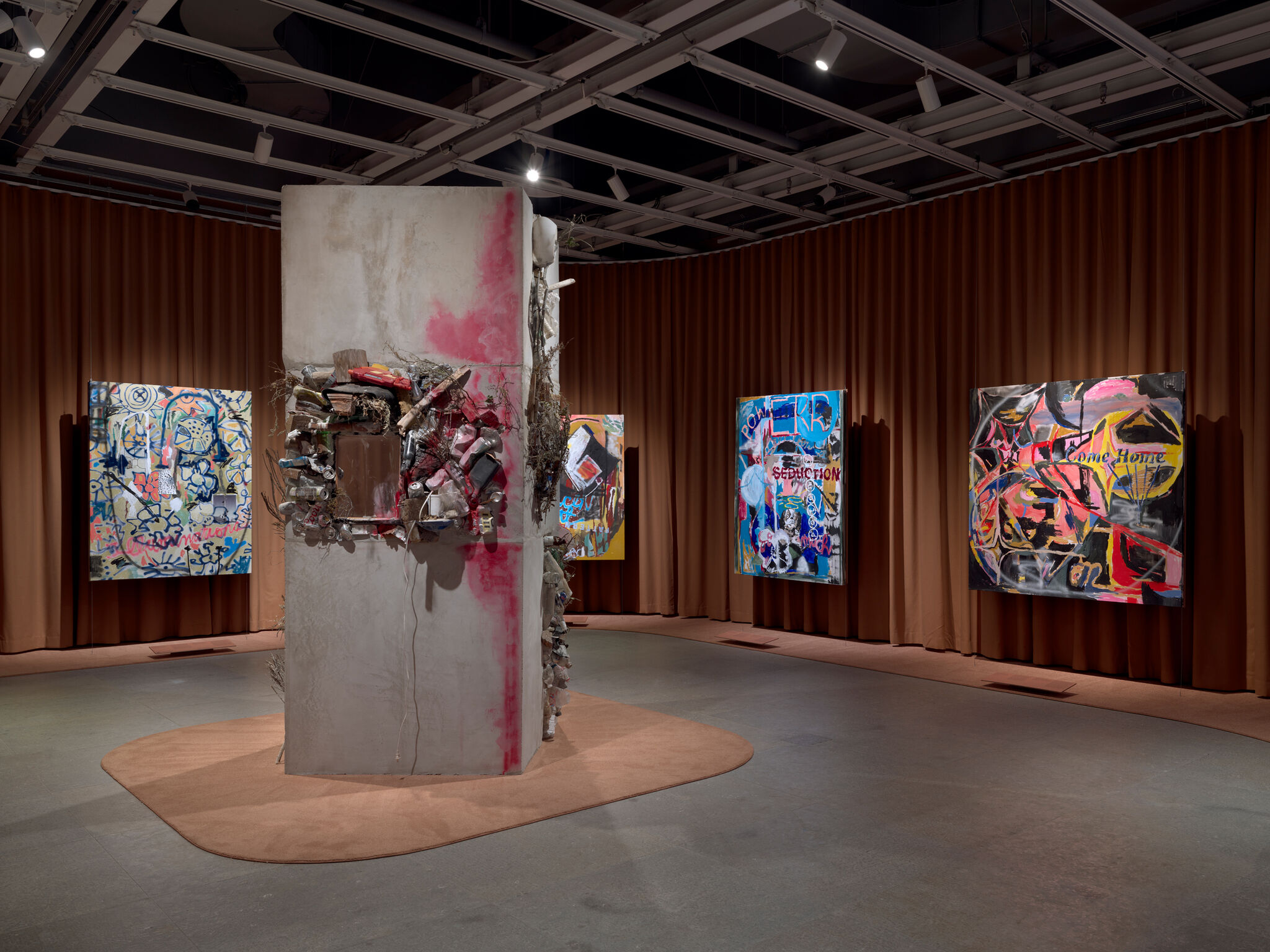Concrete pillar sculpture covered with found objects and pink paint surrounded by colorful abstract paintings.