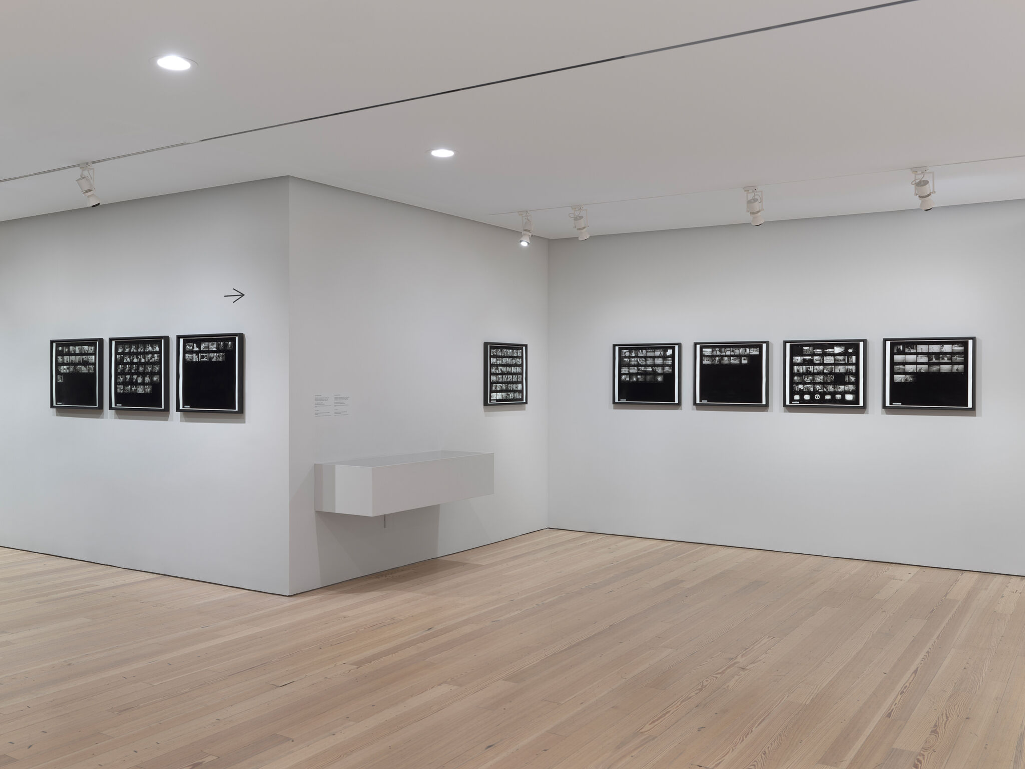 A gallery room displaying framed black-and-white contact-sheet photographs evenly spaced on pale gray walls.