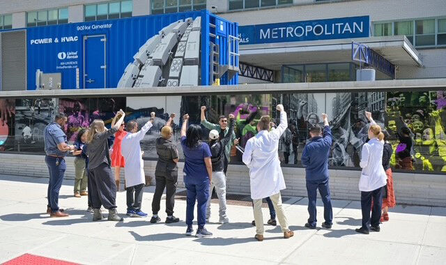 Healthcare workers and supporters stand outdoors raising fists toward a mural on a hospital building.