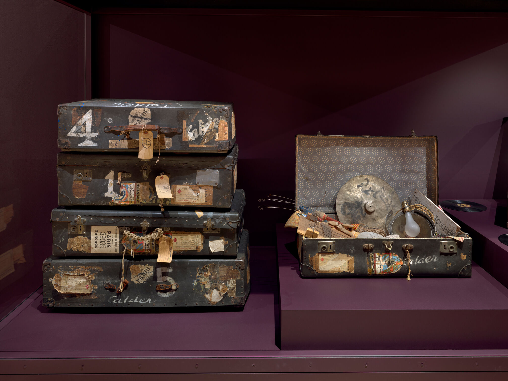 Four worn travel trunks stacked on the left and an open trunk with musical and household items on the right.