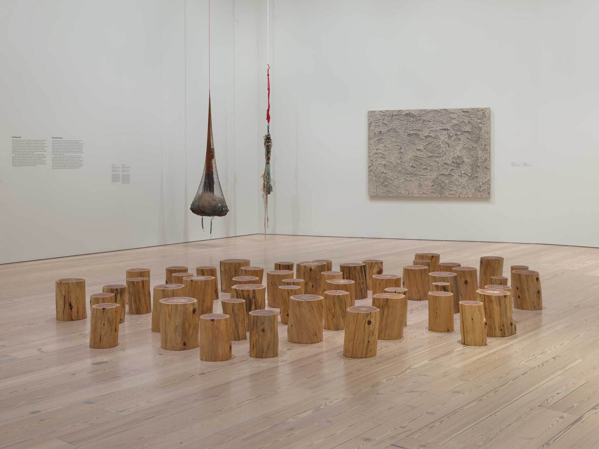 A group of wooden stump seats arranged on the gallery floor beneath hanging sculptures and a textured wall panel.