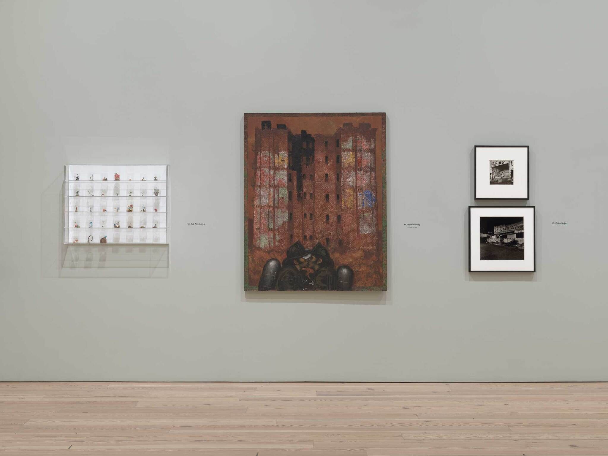 A gallery wall showing an illuminated cabinet of small objects, a large rust-colored building painting, and two black-and-white photographs.